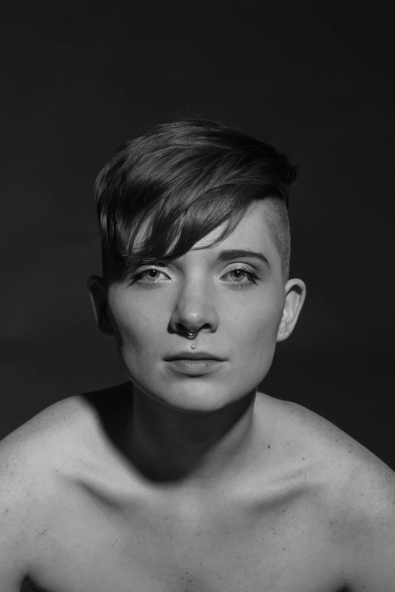 Black and white portrait of a woman with a short, styled haircut, looking directly at the camera with a neutral expression. She has a septum piercing and a labret piercing below her bottom lip, with her shoulders bare.