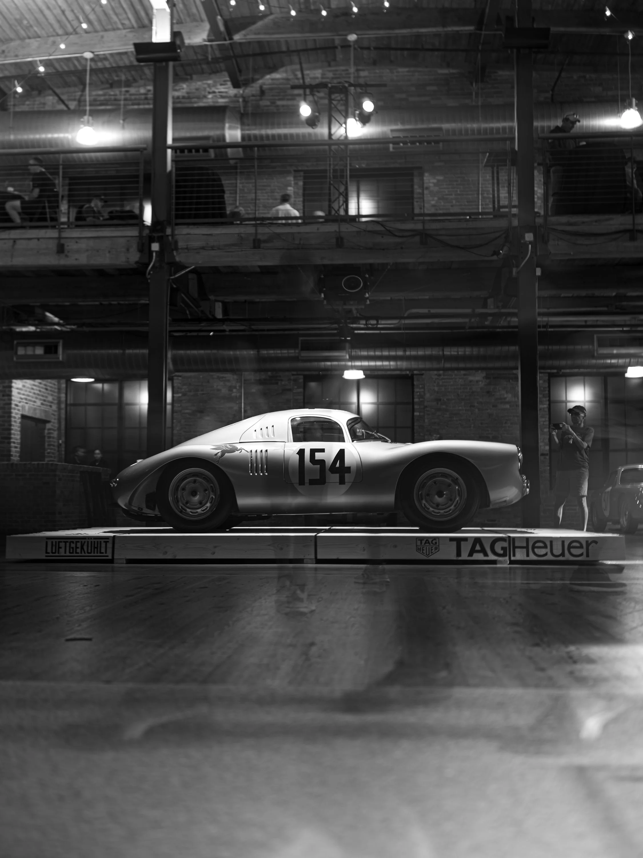 Black and white photo of a vintage race car with the number 154 on it, displayed inside a building with brick walls and wooden floors, with individuals observing from an upper level.