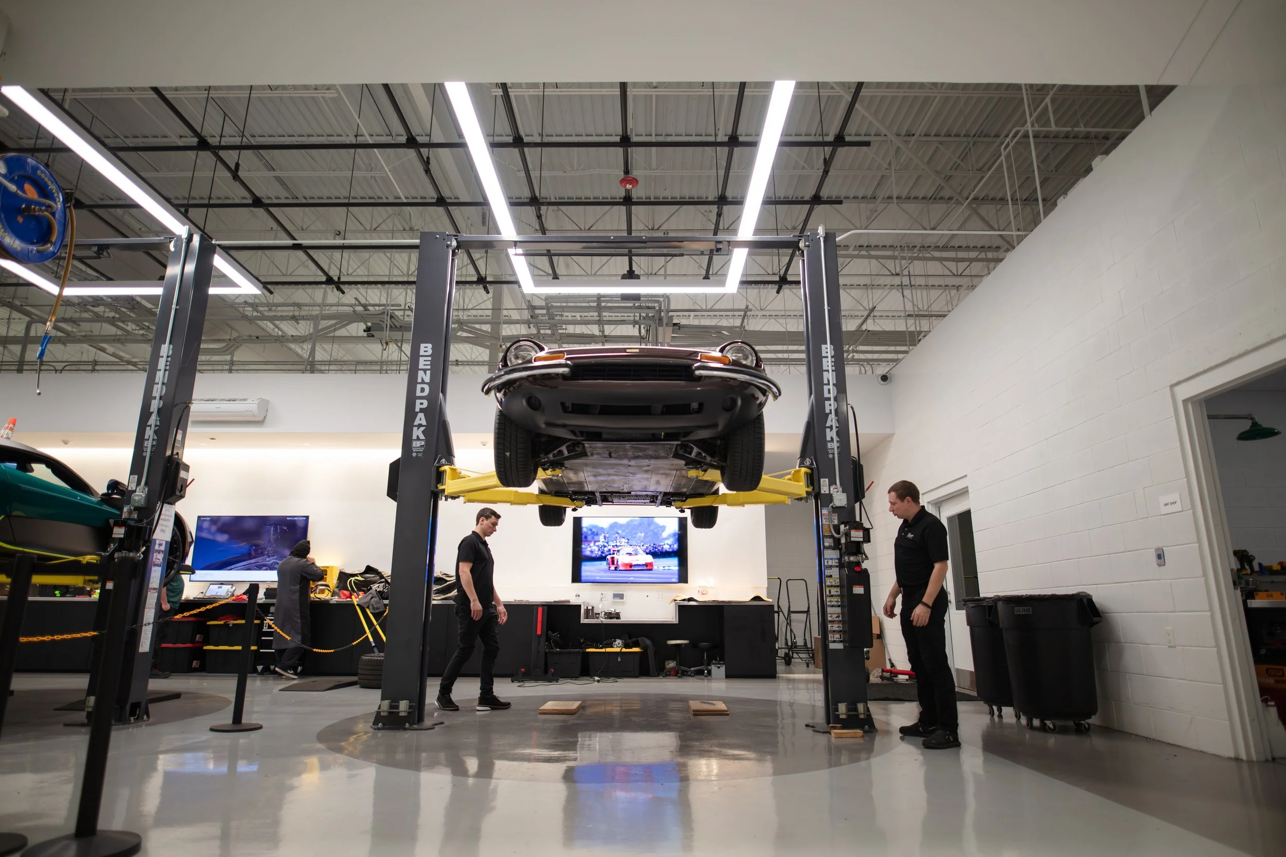 An indoor automotive workshop with a car lifted on a hydraulic lift, three people working around it, a large TV screen displaying a race car, and various equipment and tools in the background.