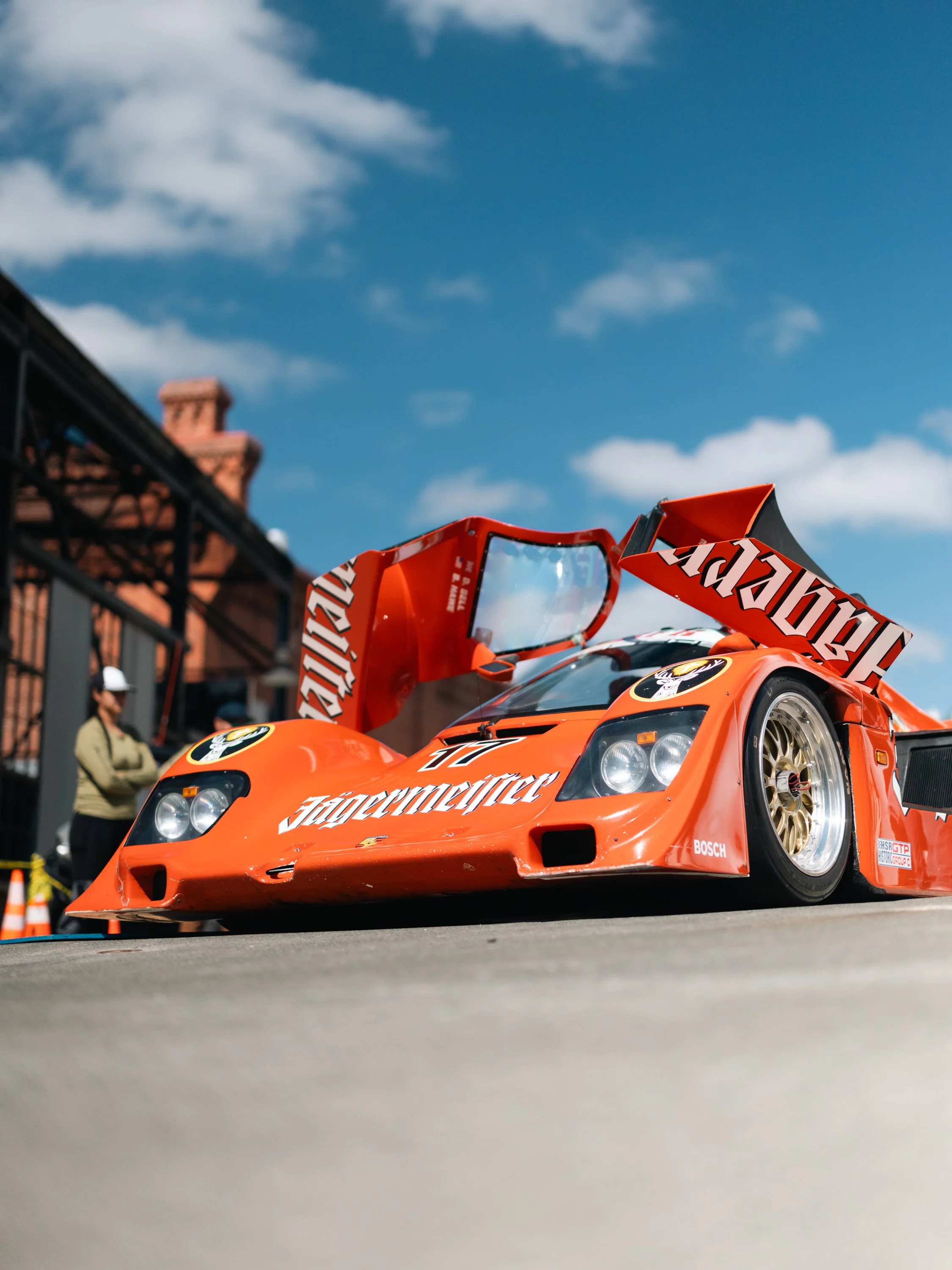 Orange racing car with sponsor logos, including Jägermeister, on a city street under blue sky with a few clouds, with a person standing nearby and buildings in the background.