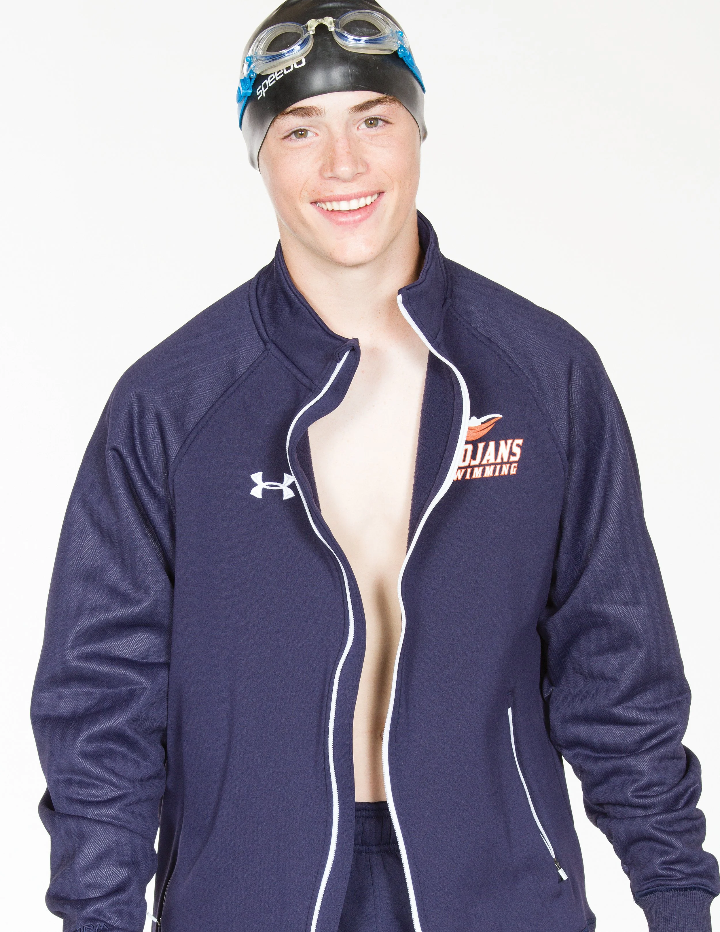 Young male swimmer wearing a black swim cap and goggles, smiling, in a navy blue zip-up jacket with team logo, against a plain white background. Under Armor.