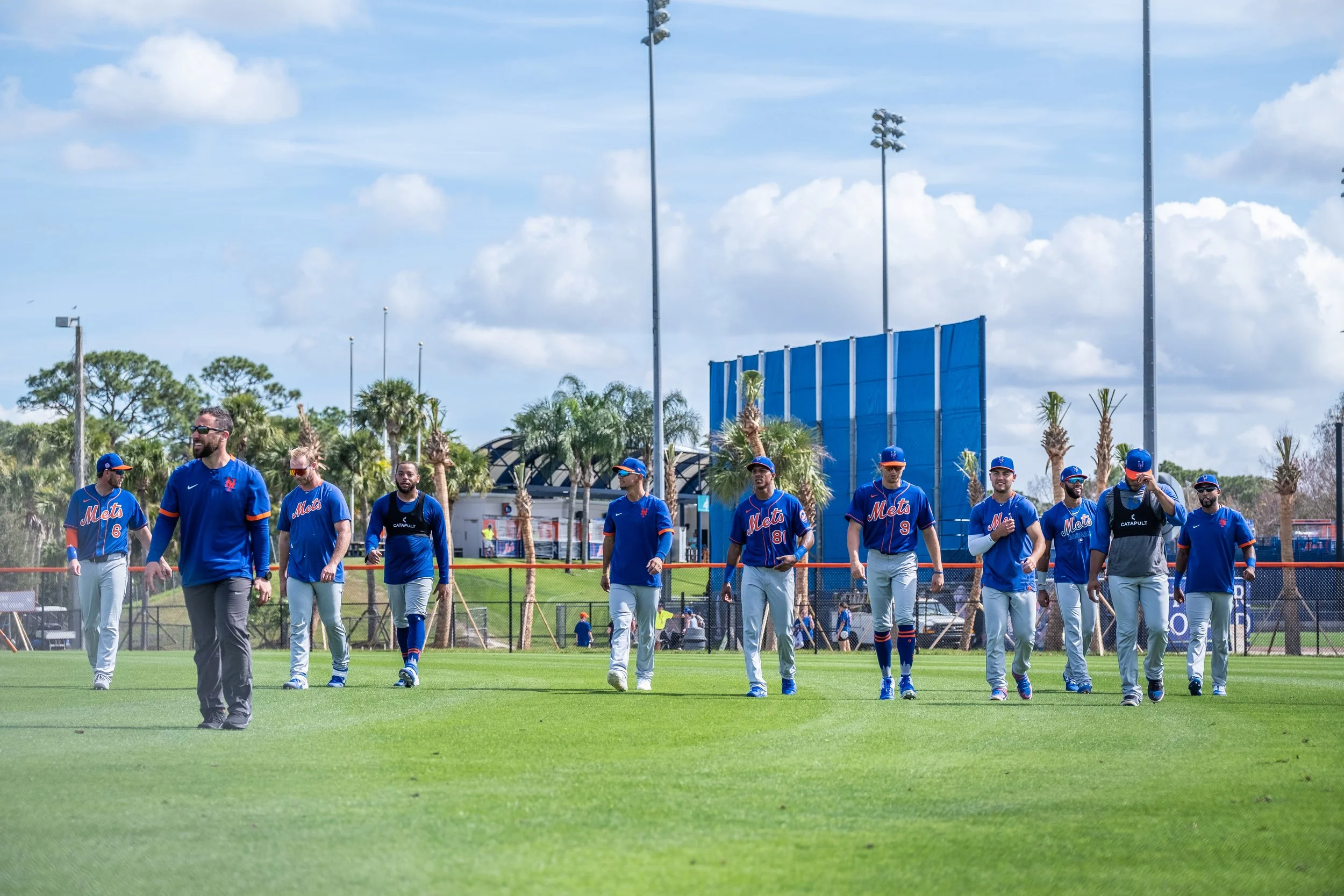 A group of baseball players wearing blue uniforms with 'Mets' written on them, walking on a lush green field under a partly cloudy sky, with palm trees and tall stadium lights in the background.