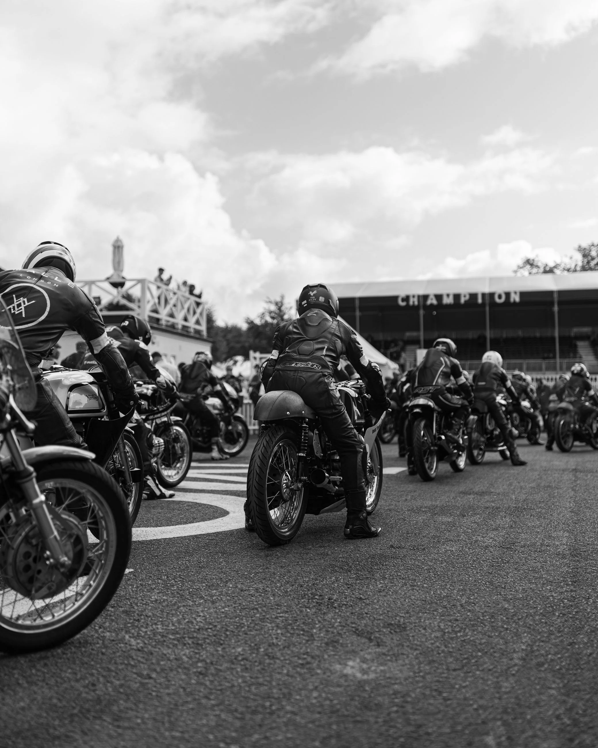Motorcycles lined up at a race track with a person in black gear sitting on one of the bikes. There are spectators and a tent with the word 'CHAMPION' in the background, under a partly cloudy sky.