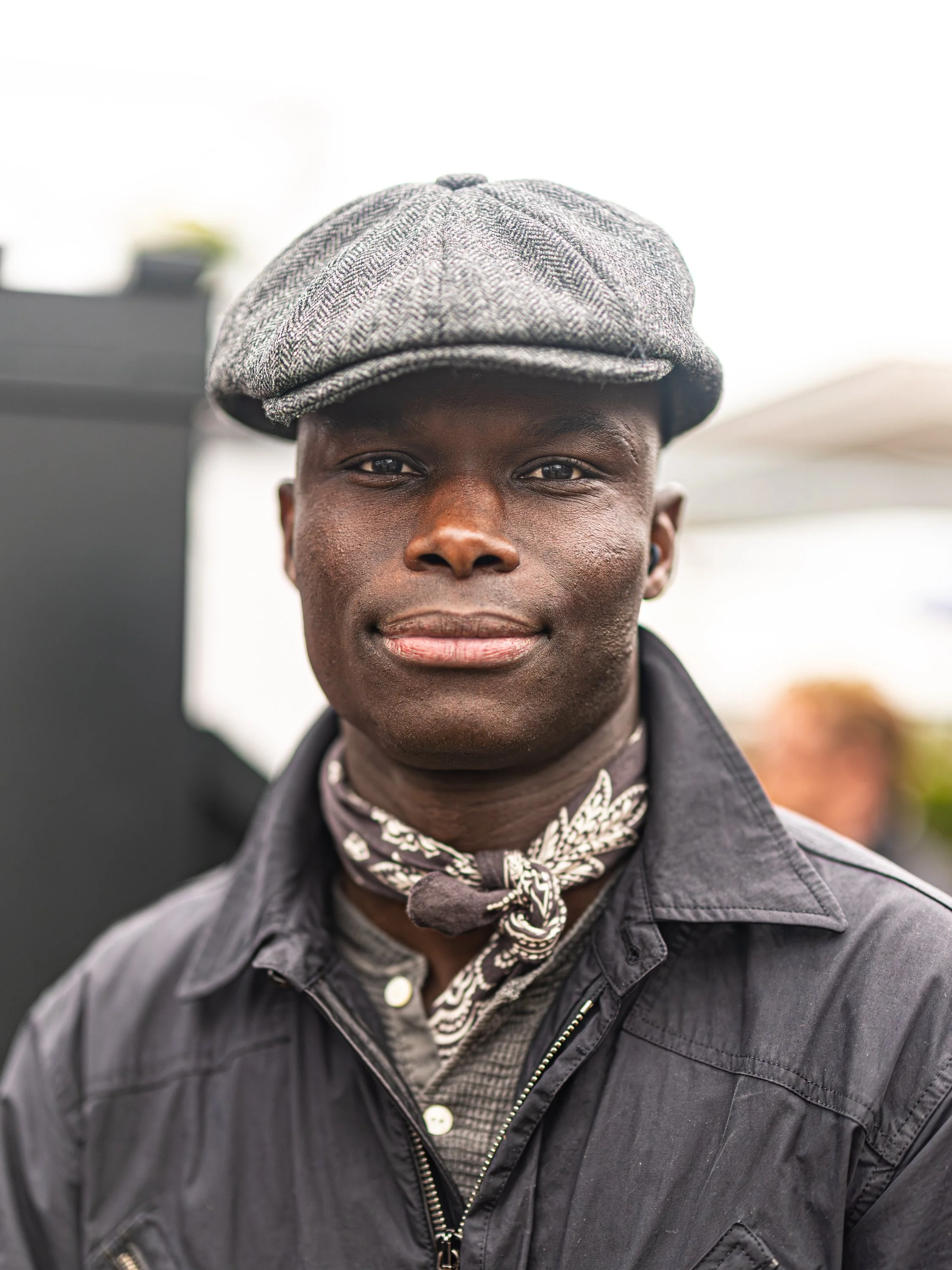 Close-up of a young man wearing a gray cap, black leather jacket, and patterned bandana around his neck, with a blurred outdoor background. Goodwood Revival.