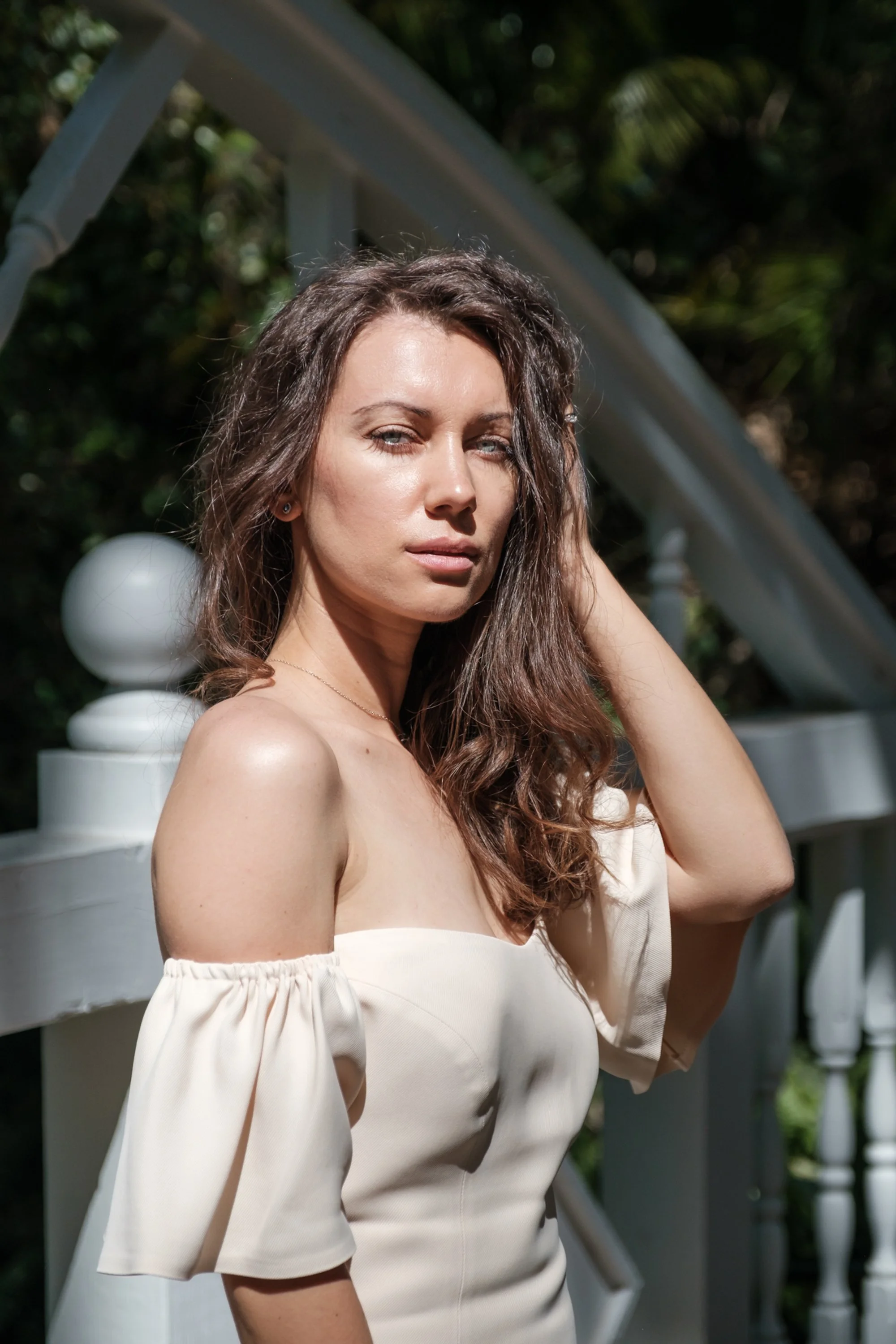 A woman with wavy brown hair and blue eyes posing outdoors near a white fence with rounded posts and greenery in the background, wearing an off-the-shoulder cream-colored dress.