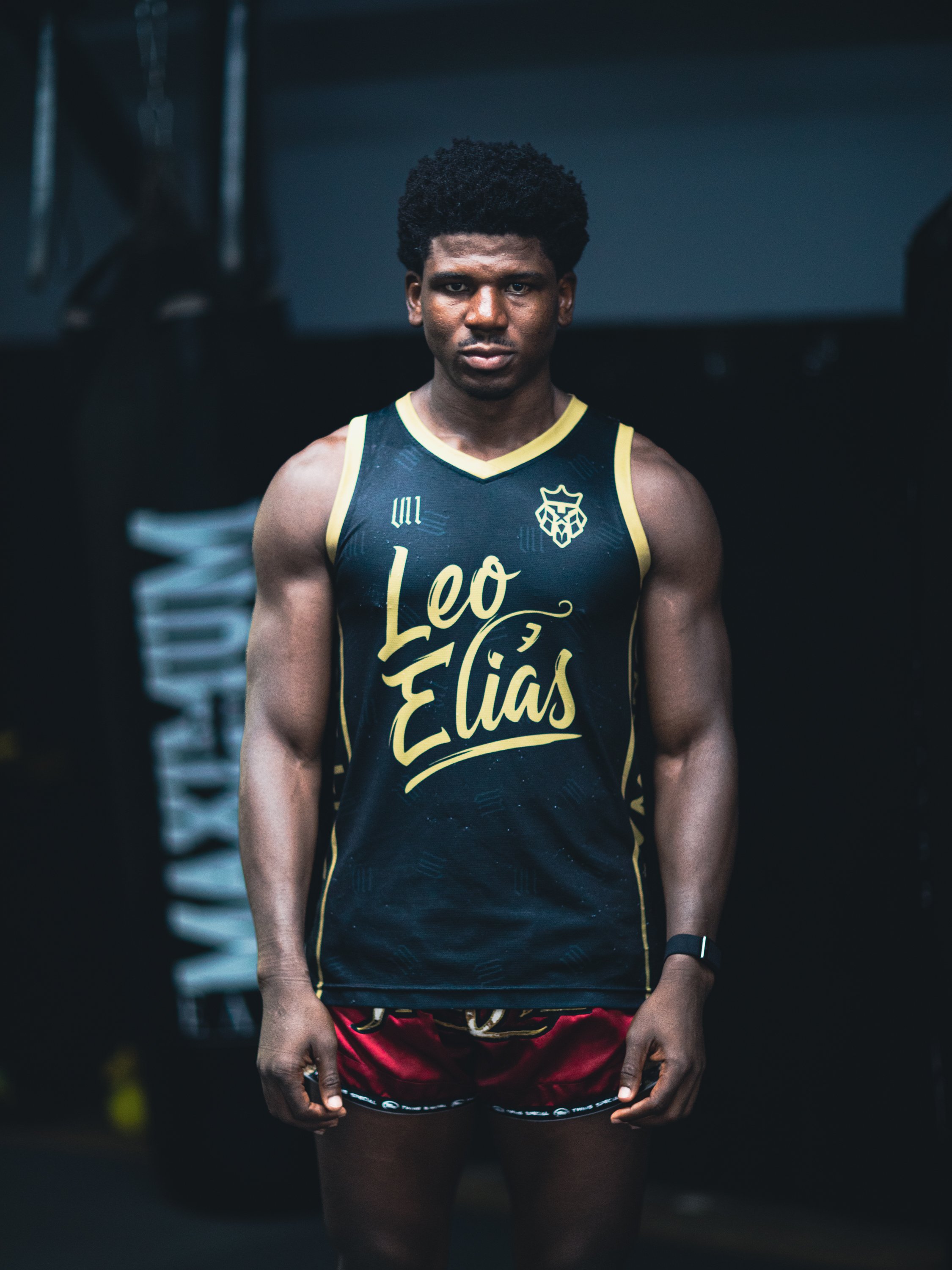 A young male athlete stands in a gym, wearing a sleeveless sports jersey with a logo and text, and red shorts, with a focused expression.