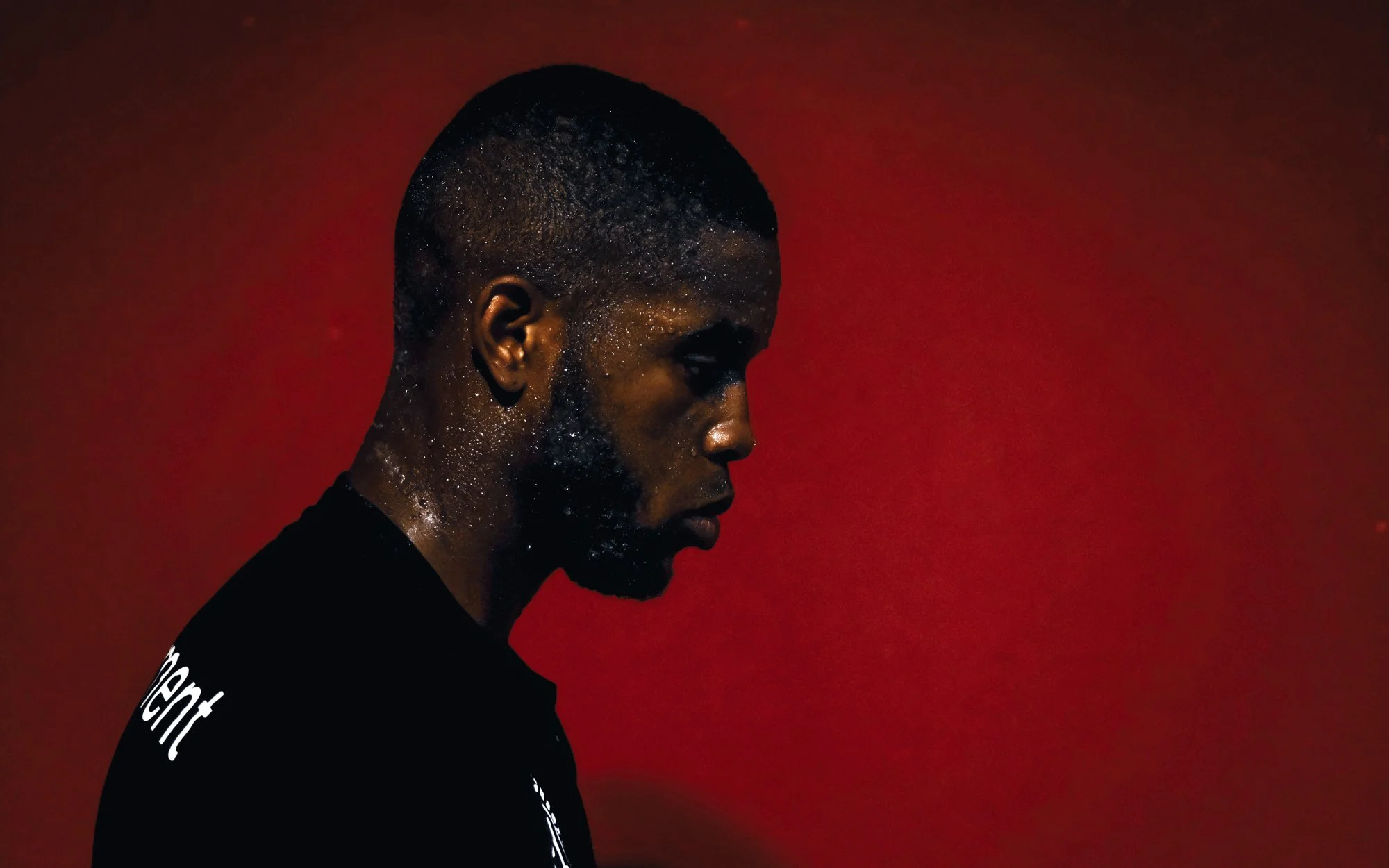 Profile of a man with dark skin, short textured hair, and a beard, looking down against a red background.