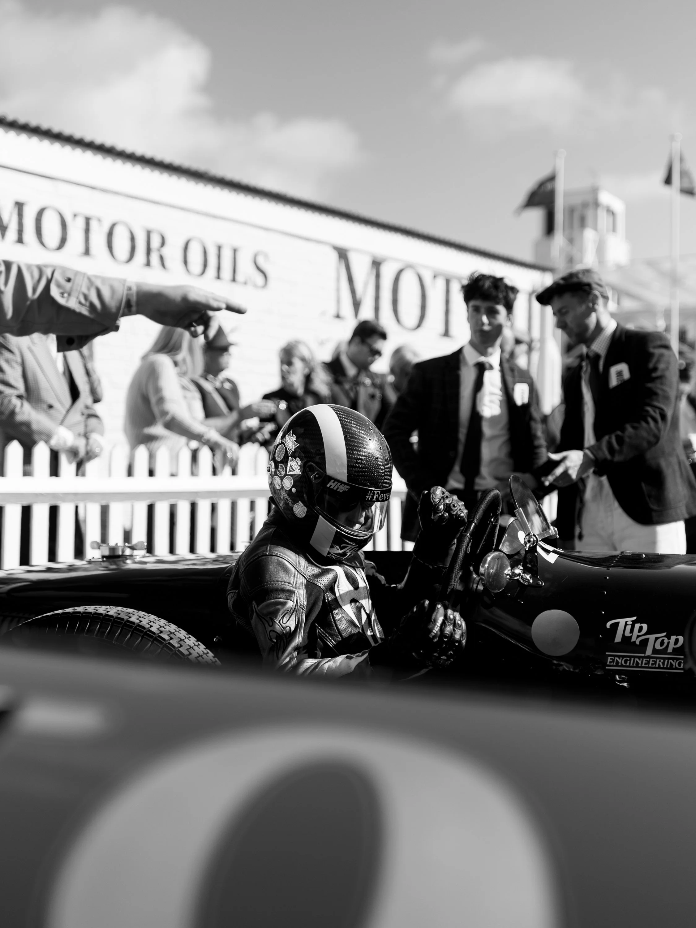 Black and white photo of a race car driver in a helmet sitting in a vintage race car, with people and a building in the background at a motorsport event.
