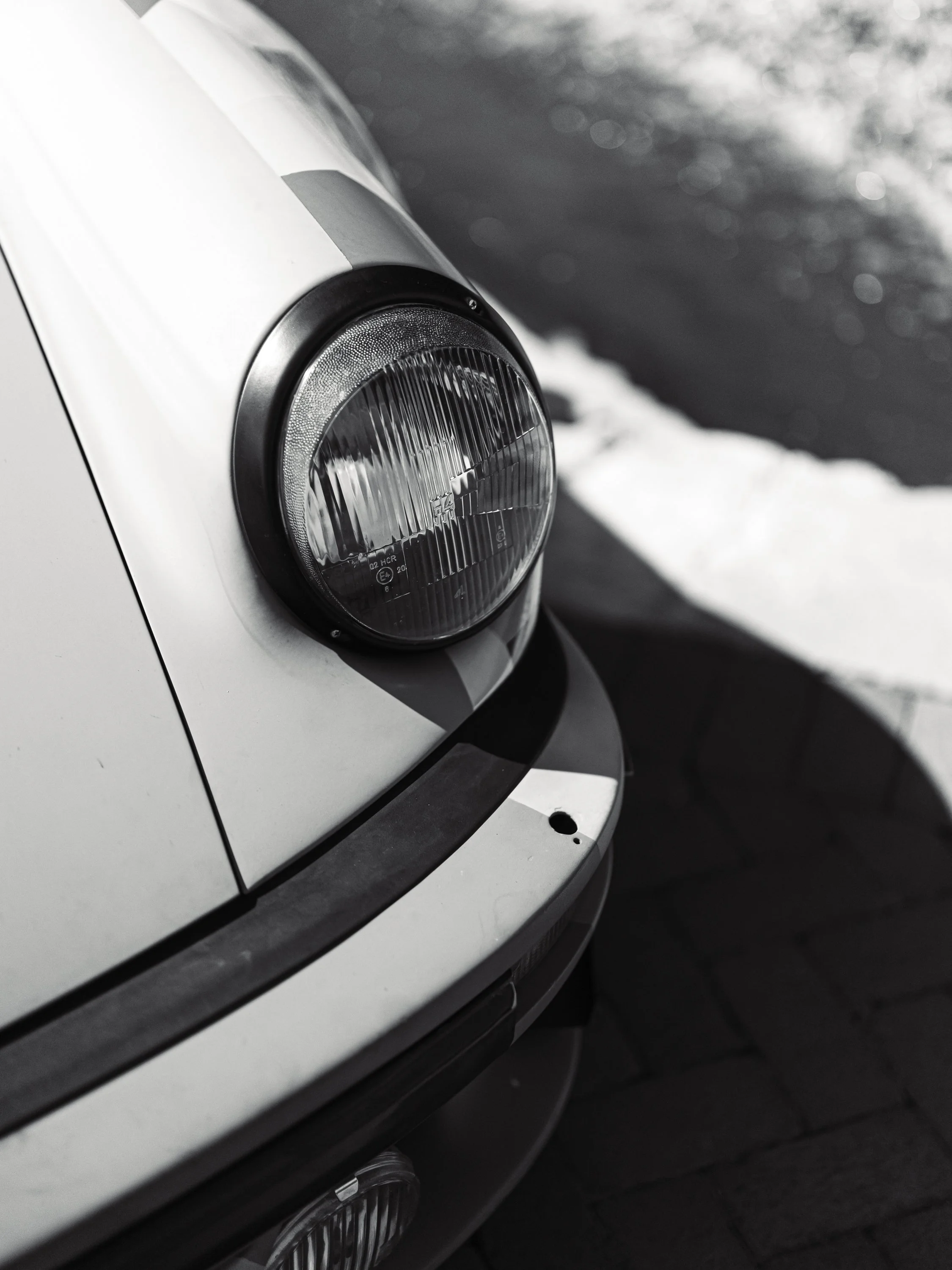 Close-up of the front right headlight of a silver car with a black bumper, parked on a dark street with snow and ice.