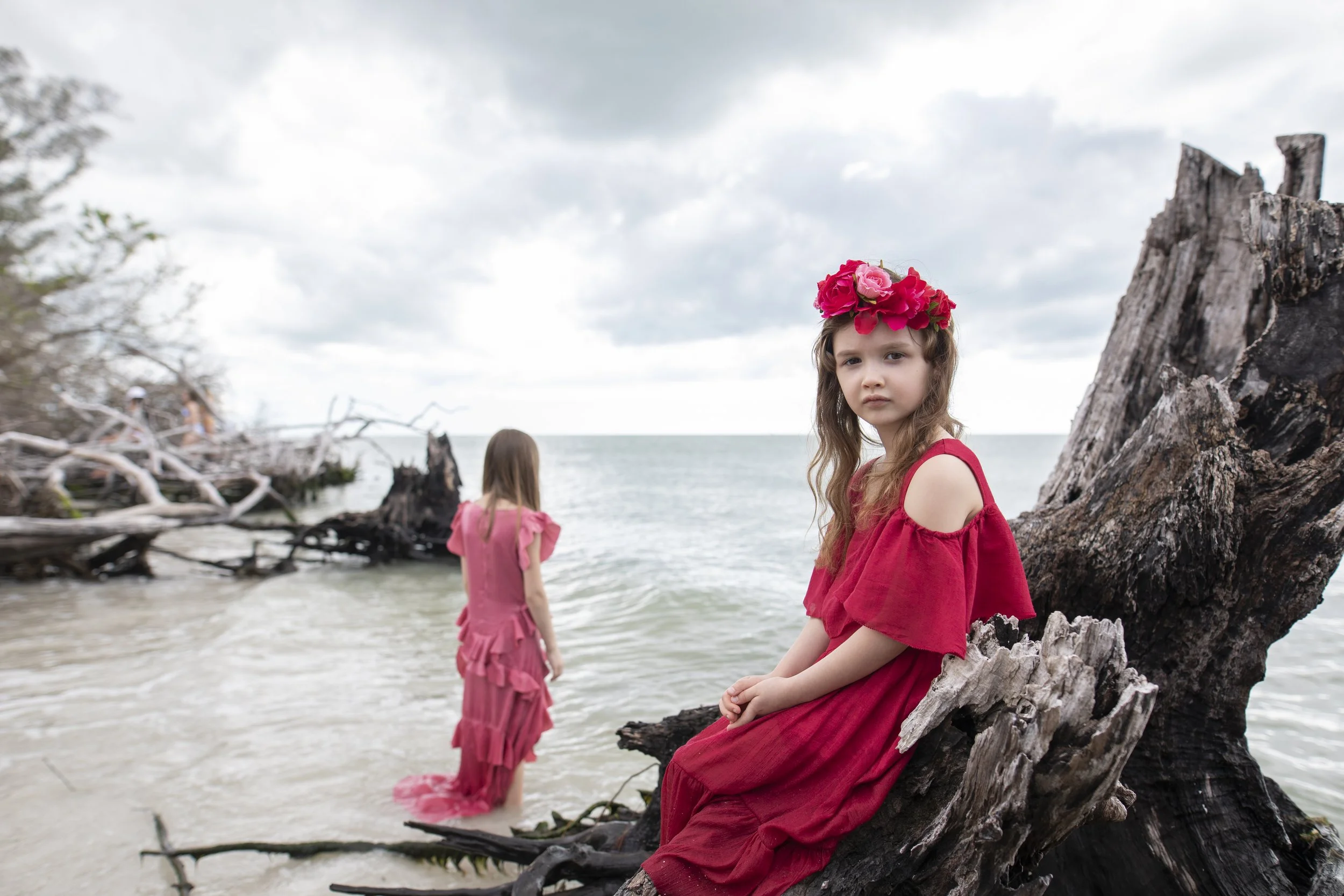 Three girls in red dresses at the beach, with one girl sitting on a driftwood log wearing a floral crown, and two other girls standing near the water, with cloudy skies overhead.