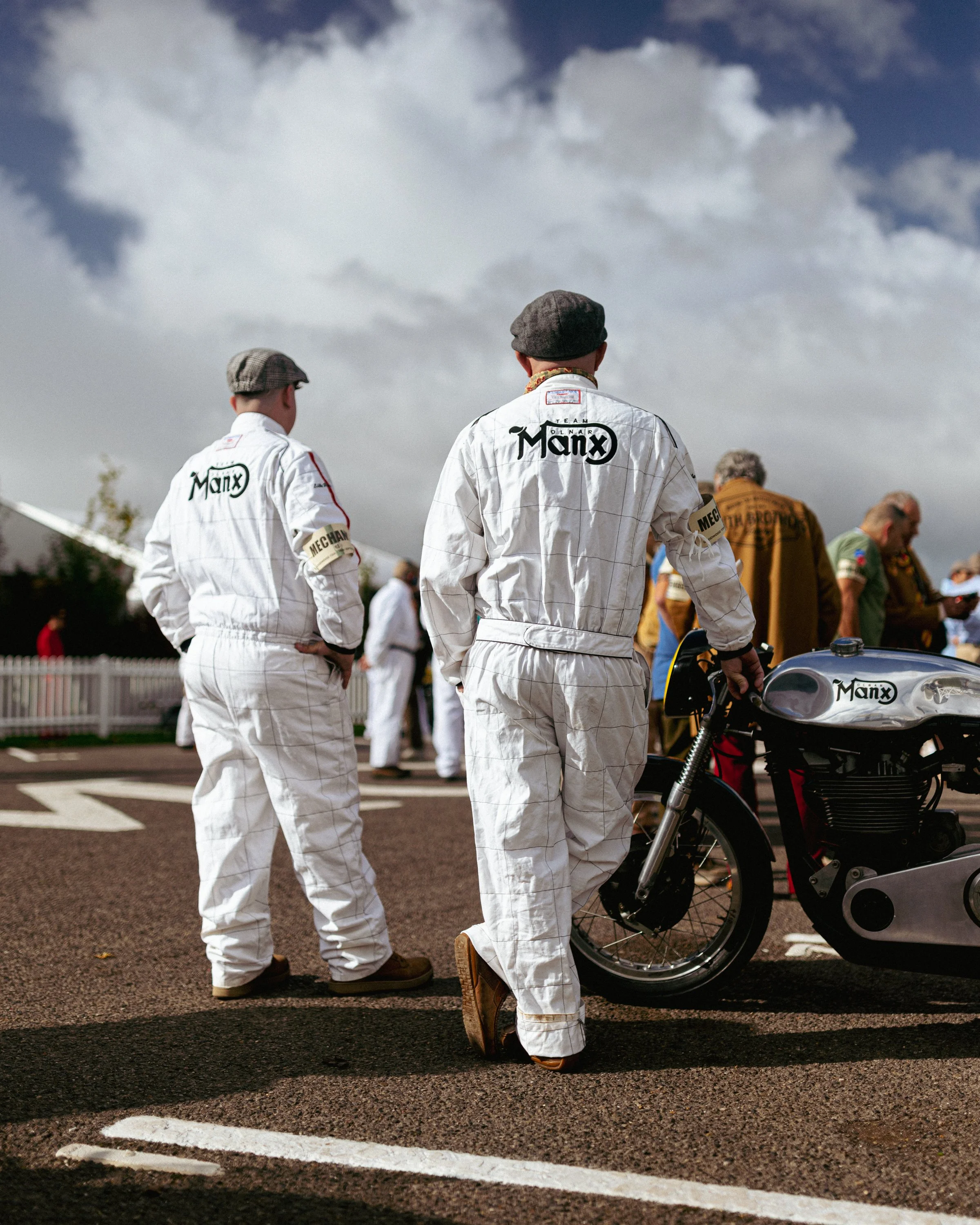 Two men dressed in white racing suits and tweed flat caps standing near a motorcycle with a fuel tank inscribed 'Manx' during daytime at a racing event with other people in the background under cloudy skies.