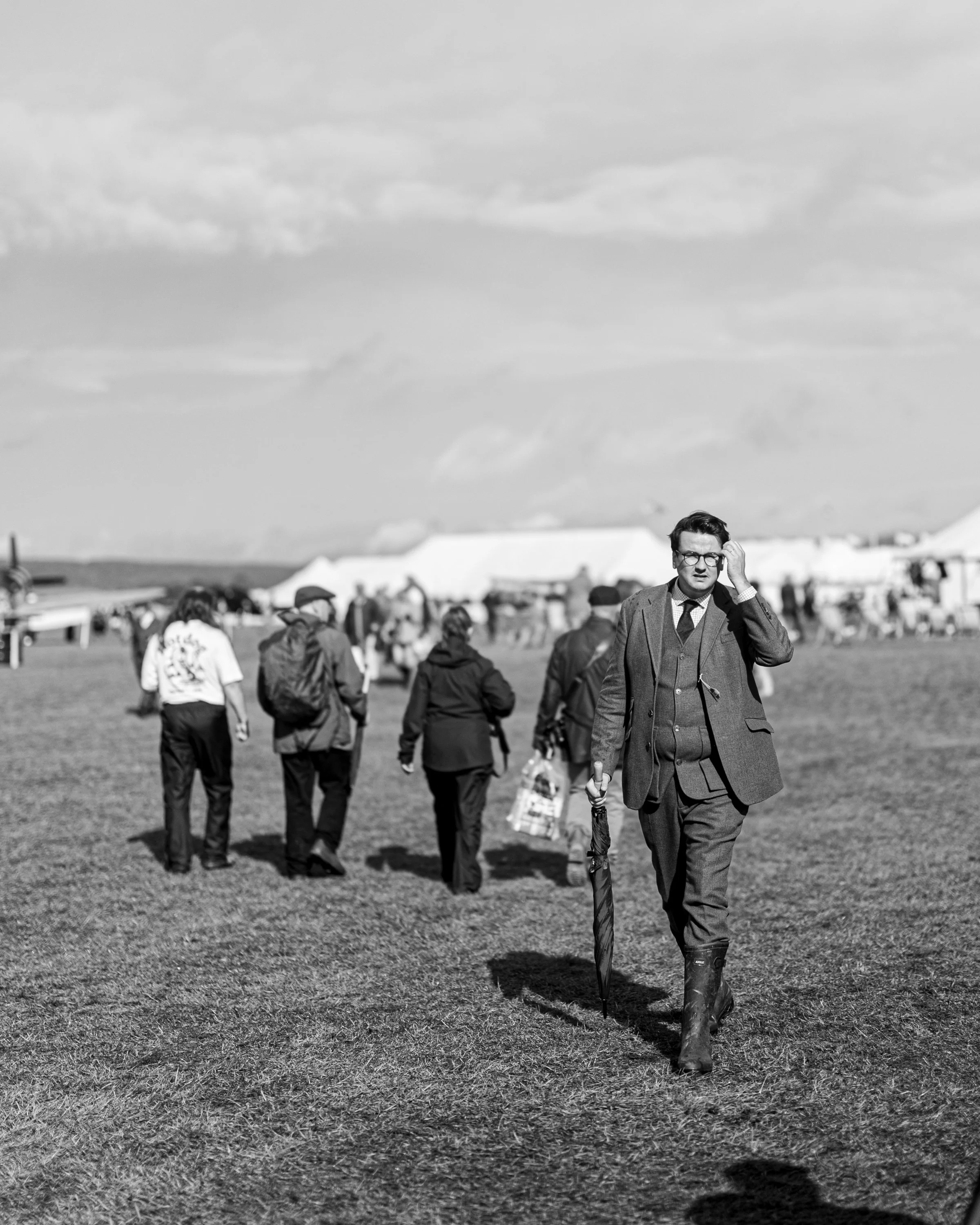 A man in a suit and glasses walking on a grassy field with an umbrella, under a cloudy sky, with several people in the background.