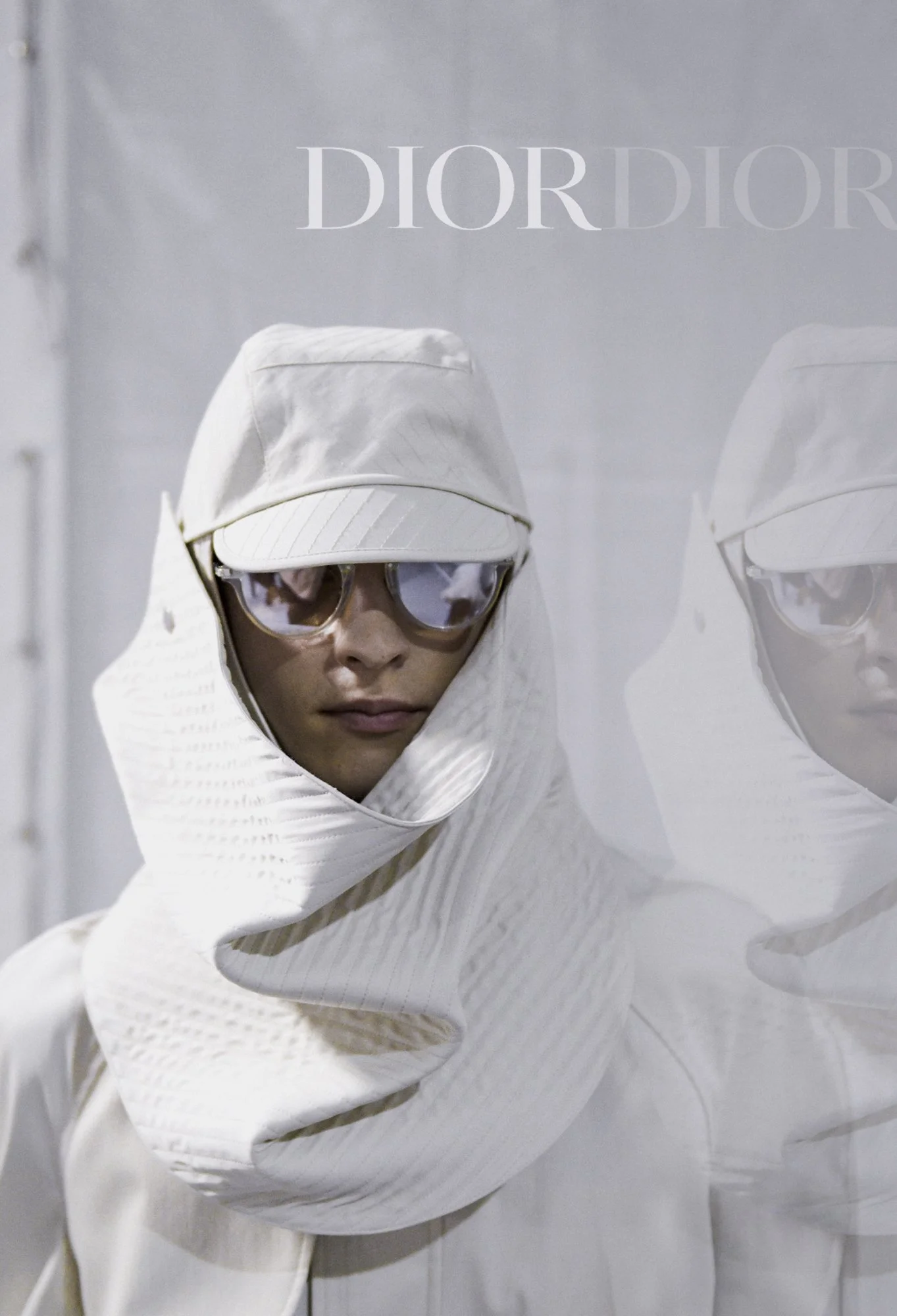 A person dressed in a white hooded outfit and reflective sunglasses, standing in front of a white background with the word DIOR written on it.