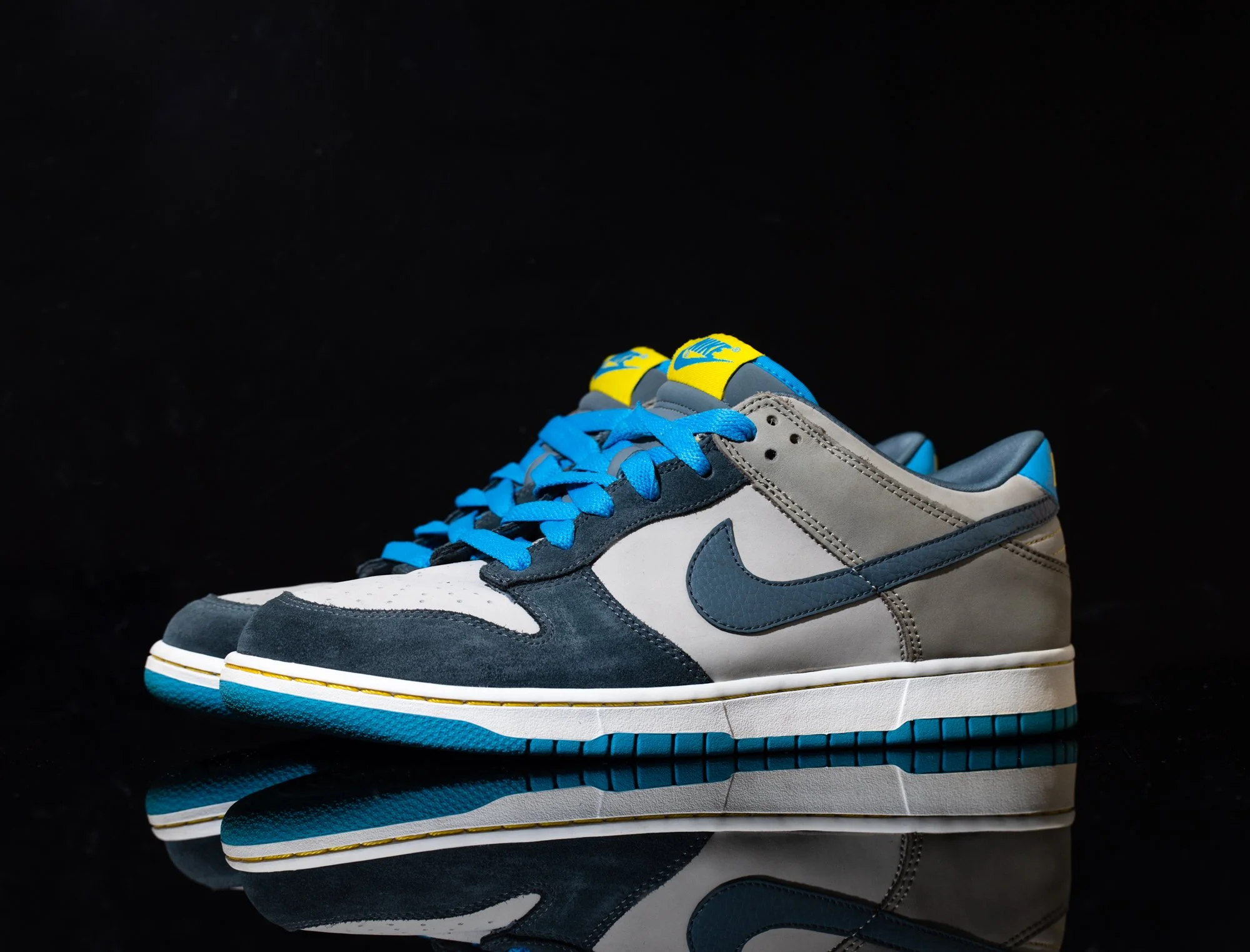 A pair of Nike sneakers with a combination of grey, white, black, and blue colors, featuring blue laces and a yellow detail on the tongue, placed on a reflective black surface. Nike SB.