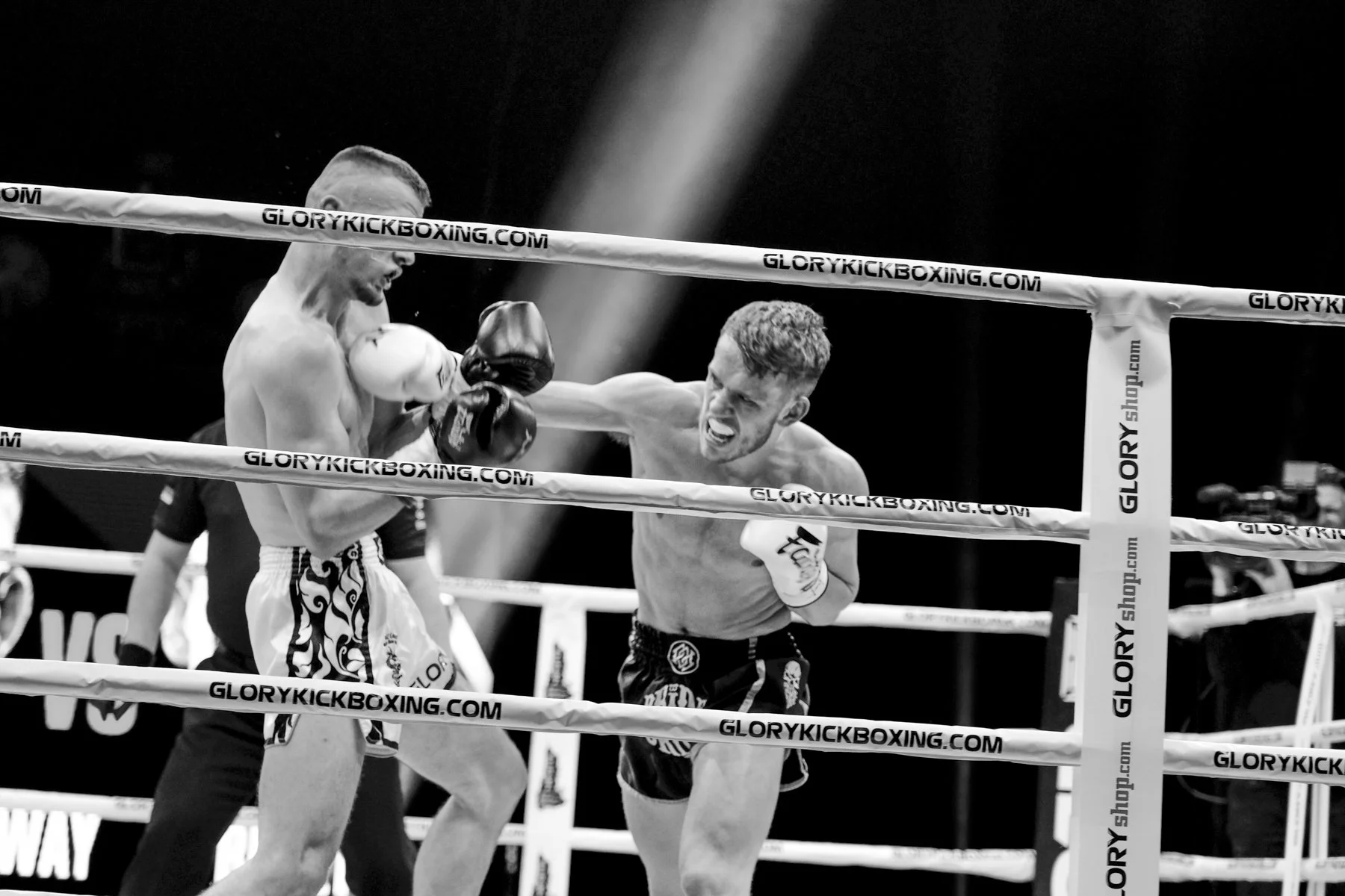 Black and white photo of two male kickboxers in a ring engaged in combat. The fighter on the left is throwing a punch, while the fighter on the right is blocking or reacting to the punch. The ring has banners with the text 'GLORYKICKBOXING.COM'. Ther