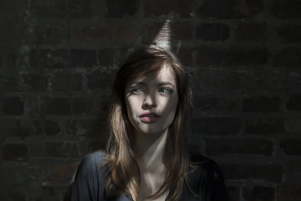 A young woman with long brown hair and blue eyes looks to her left. She is standing against a dark brick wall with a small beam of light shining in from above, illuminating part of her face.