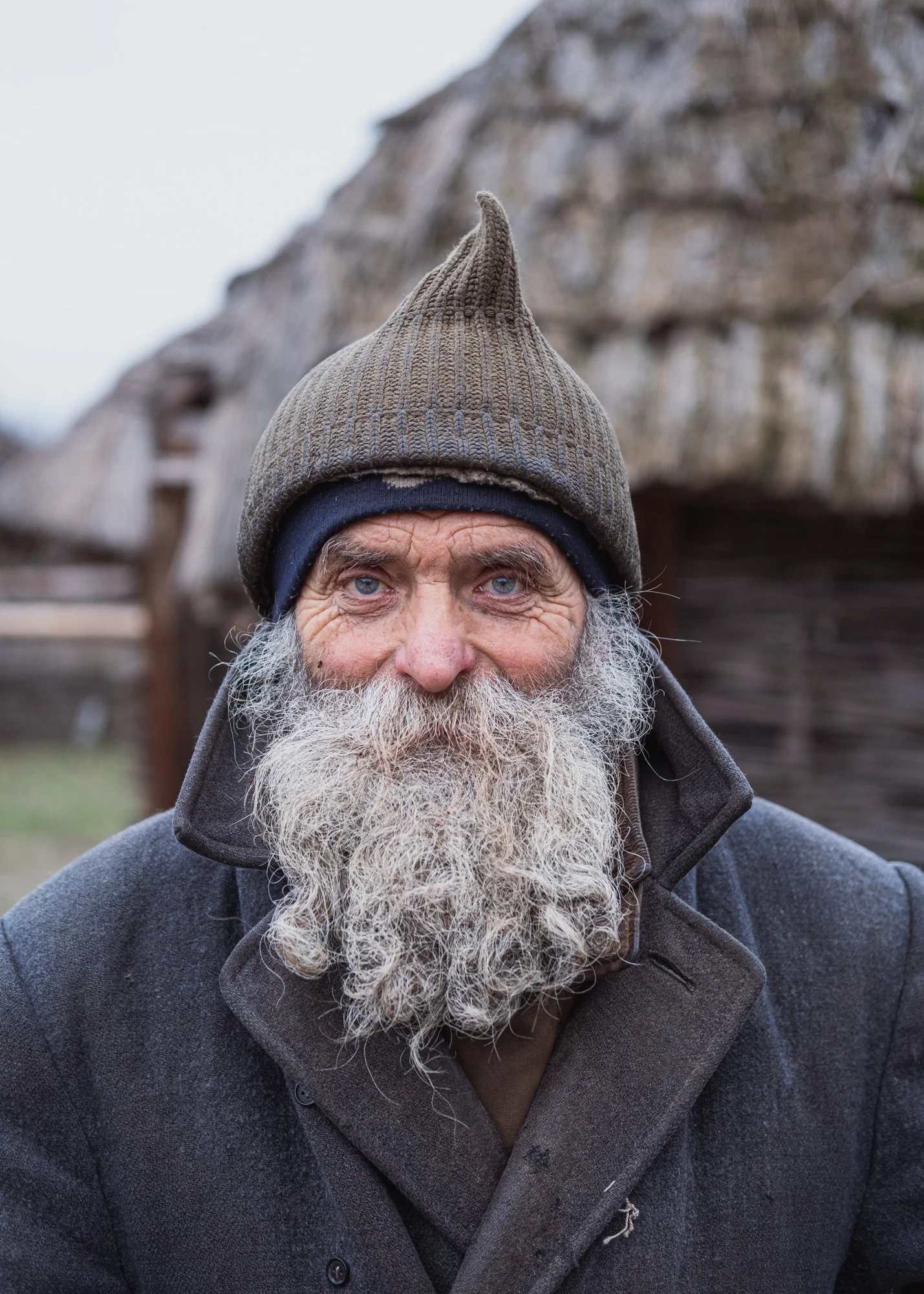 Close-up of an elderly man with a long beard and blue eyes, wearing a brown knit hat with a pointed top and a dark gray coat, outdoors with blurred wooden structures in the background. Ukraine.