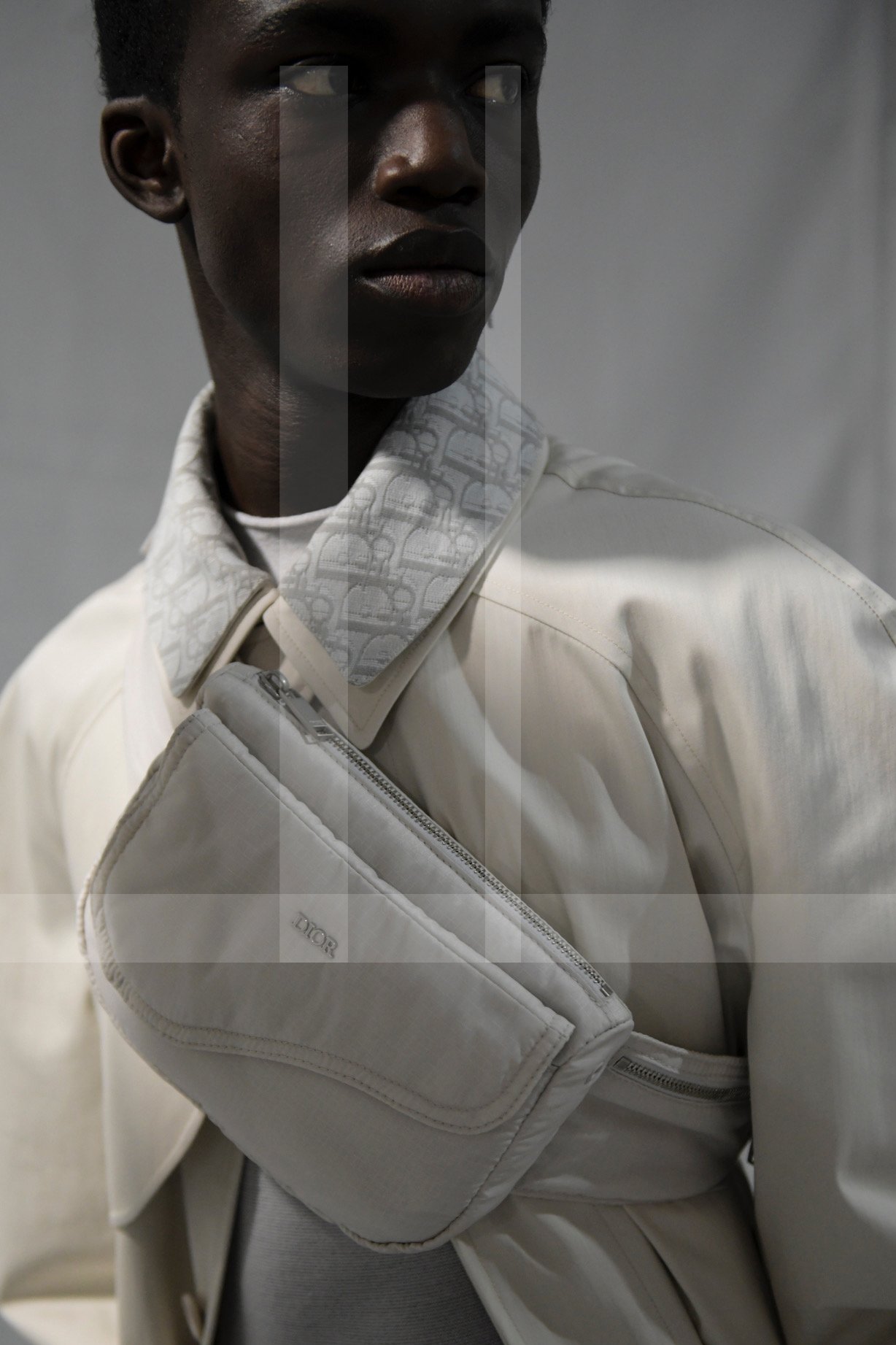 A person wearing a white Dior bag crossbody, a patterned collar shirt, and a cream-colored jacket, standing against a plain gray background.