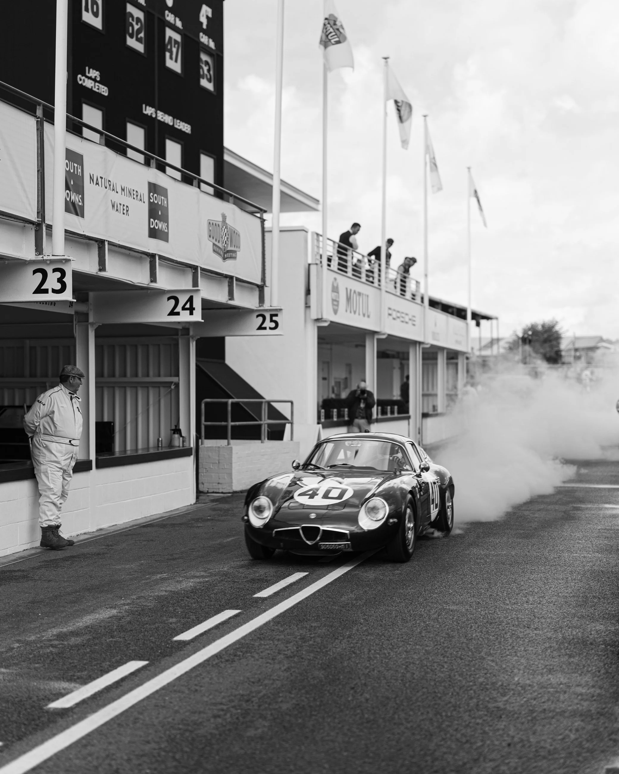 Black race car with the number 40 on the hood in the pit lane of a racing circuit, with team members and officials nearby, and a large scoreboard in the background.