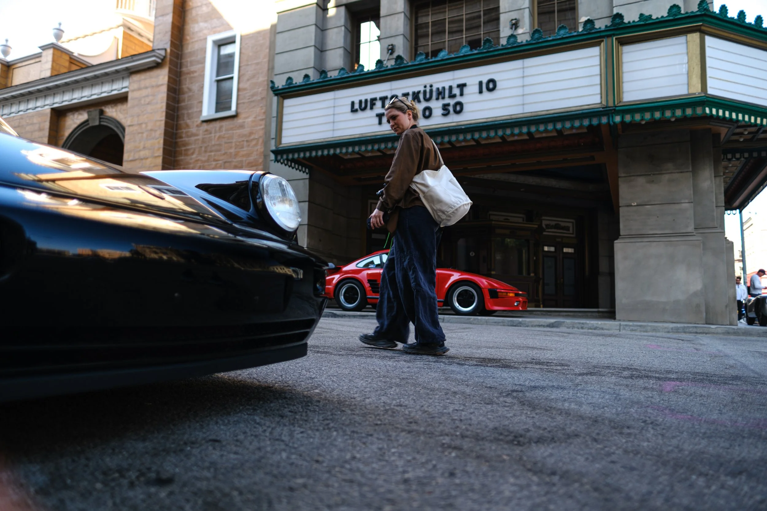 A woman with a white bag standing on a city street near a black sports car and a red sports car, with a marquee sign and historic building in the background.