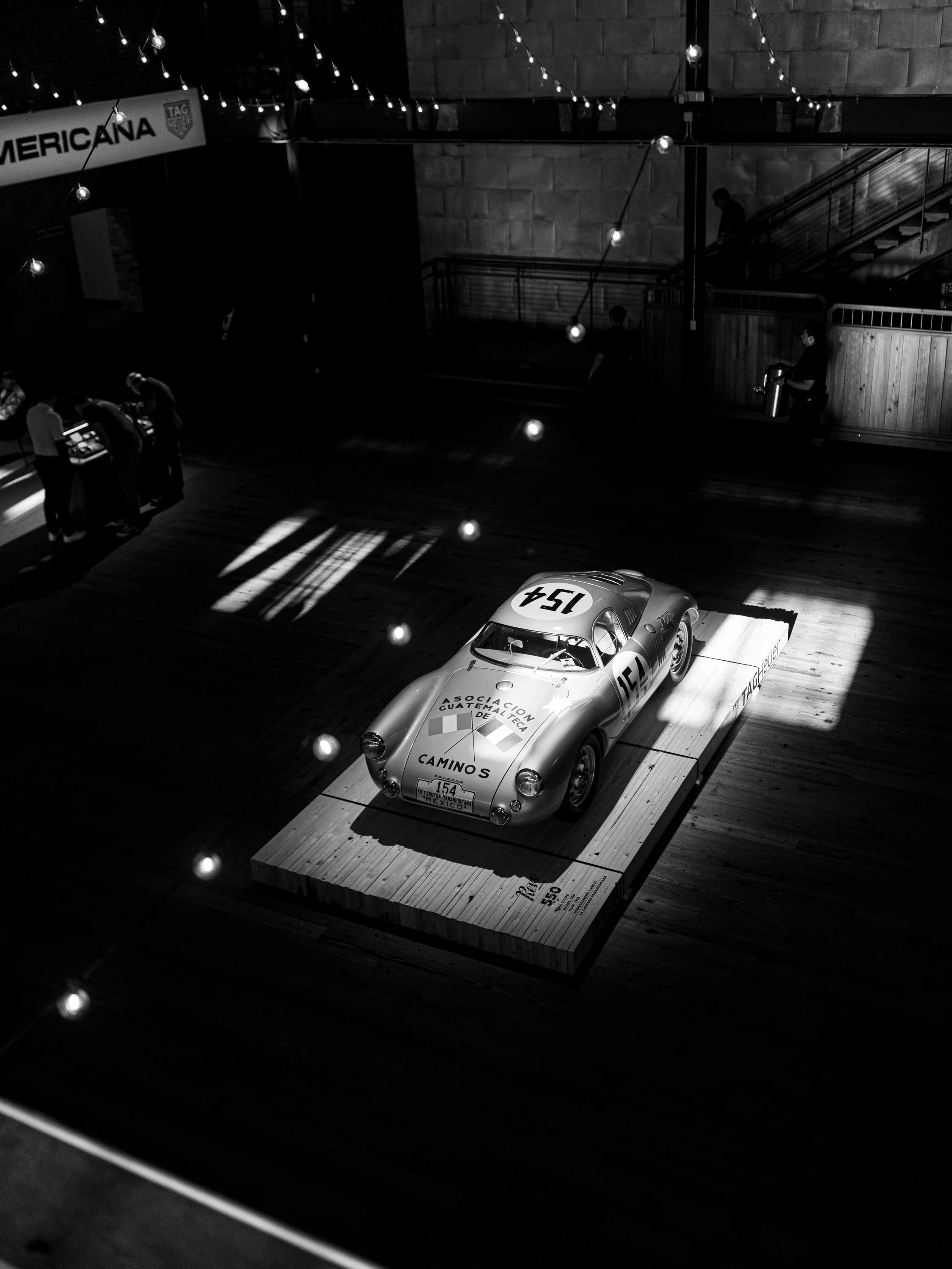 A vintage race car on display at a museum or exhibit, with labels and a platform beneath it, and people observing around.