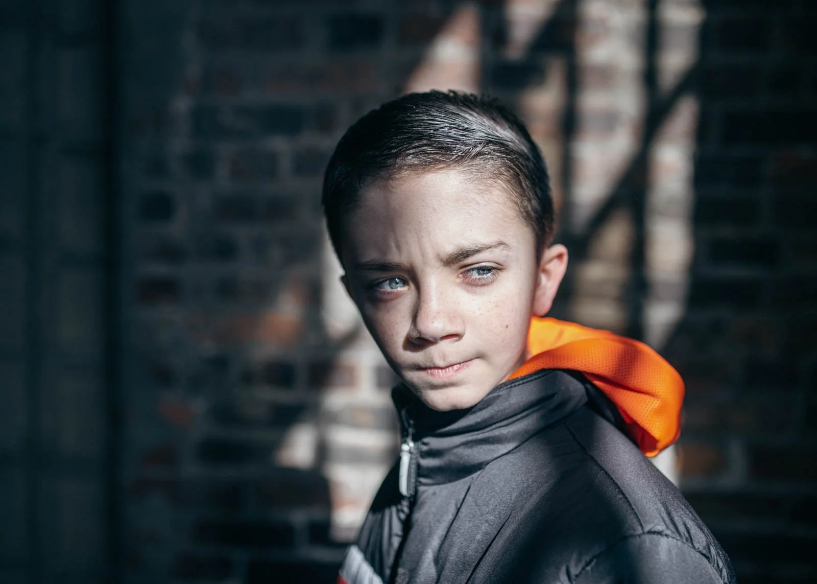 Close-up of a young boy with blue eyes, wet dark hair, wearing a black jacket with an orange hood, standing outdoors with a brick wall and fire escape stairs in the background.
