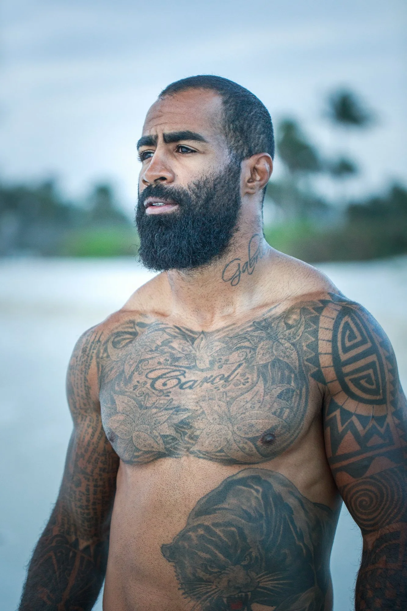 A shirtless man with a beard and tattoos standing outdoors near water with a blurred background of palm trees. Cosmo Alexandre, muay thai