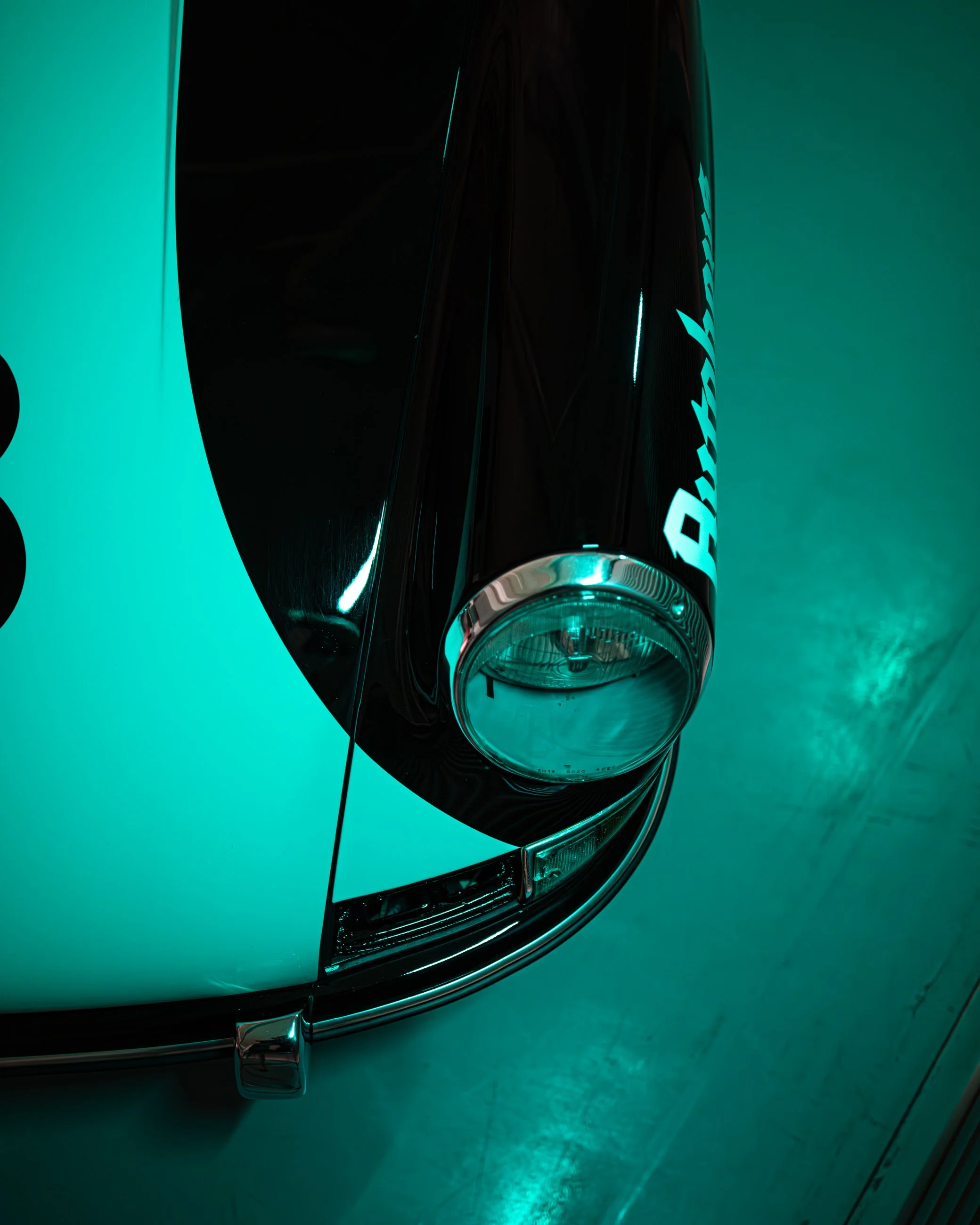 Close-up of a black bottle with a shiny cap, reflecting teal-colored light, with partial word "Bape" on the bottle, placed on a reflective surface with teal background. Porsche 911.