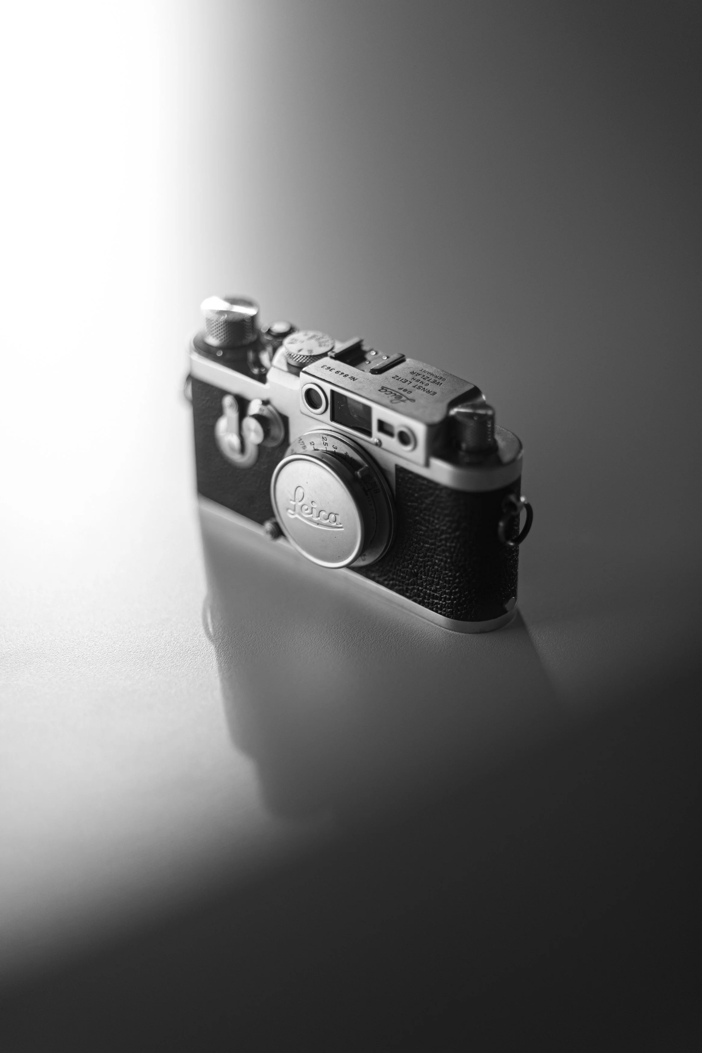 Black and white vintage Leica camera with textured grip, positioned on a surface casting a shadow, against a gradient background.
