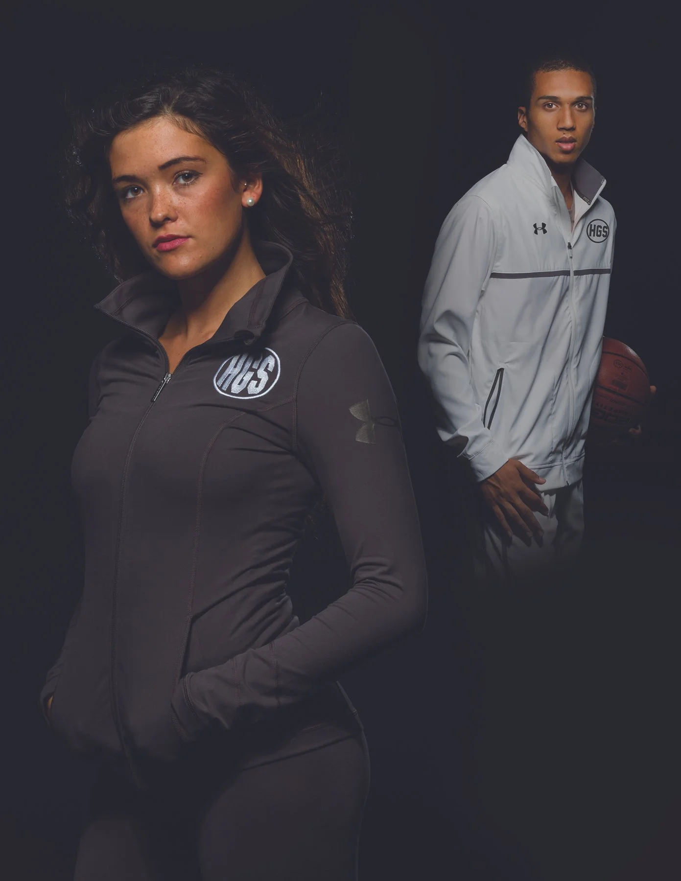 Two young athletes in athletic gear against a black background. The woman on the left has curly hair, earrings, and is dressed in a black athletic jacket with a logo. The man on the right is holding a basketball, has short hair, and is wearing a whit