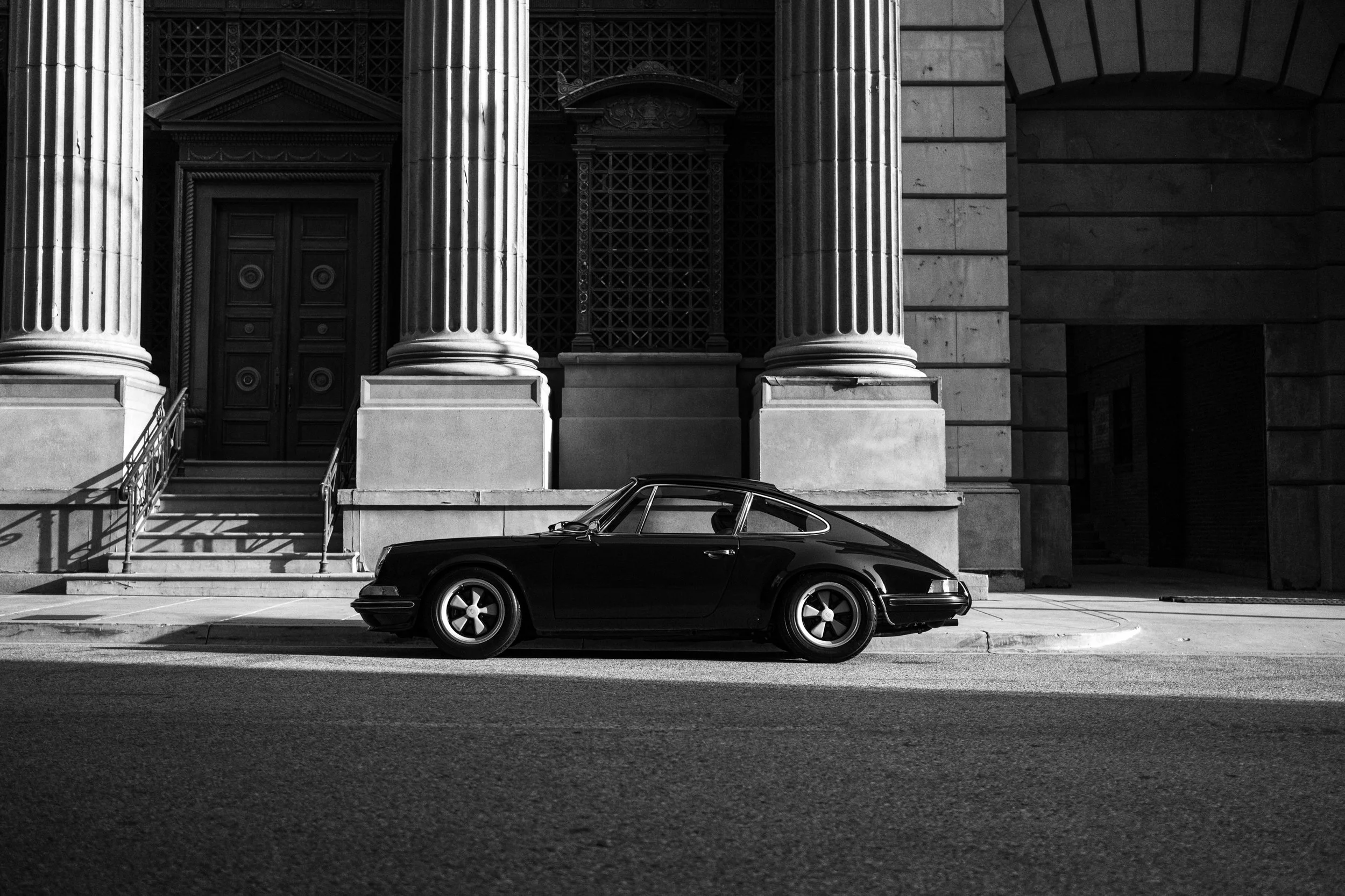 A black classic sports car parked in front of a grand, historic building with large stone columns and detailed architectural features, in black and white.