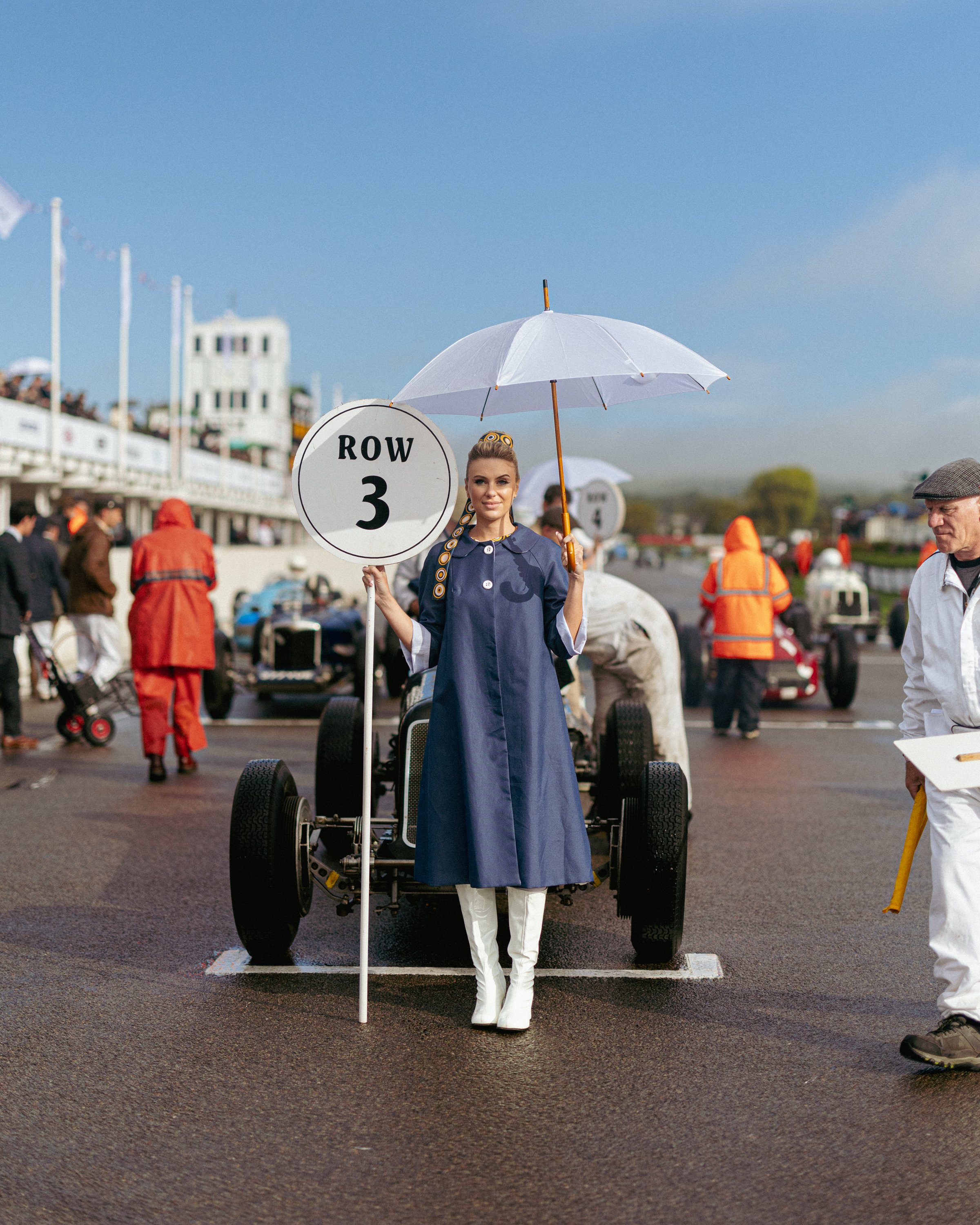 A woman dressed in vintage clothing holding a sign that reads 'Row 3' and standing in front of a race car at a racetrack, with spectators and other participants in the background.