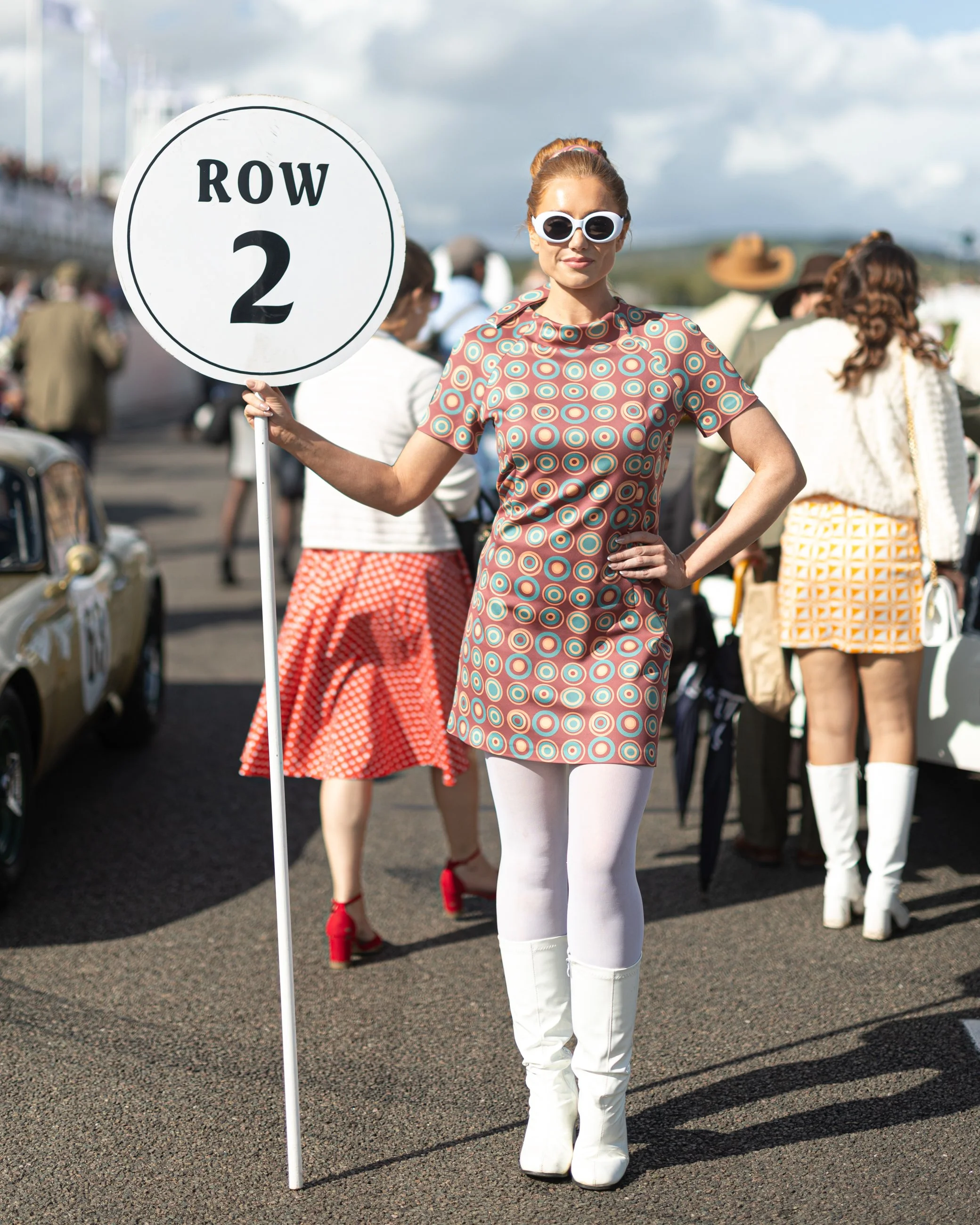 A woman dressed in retro 1960s style clothing stands next to a street sign that reads 'ROW 2'. She is wearing large white sunglasses, a patterned dress with circles, white tights, and white platform boots. She has her right hand on her hip and is sta