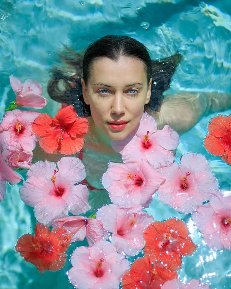 A woman with wet dark hair floating in water, surrounded by pink and red hibiscus flowers.
