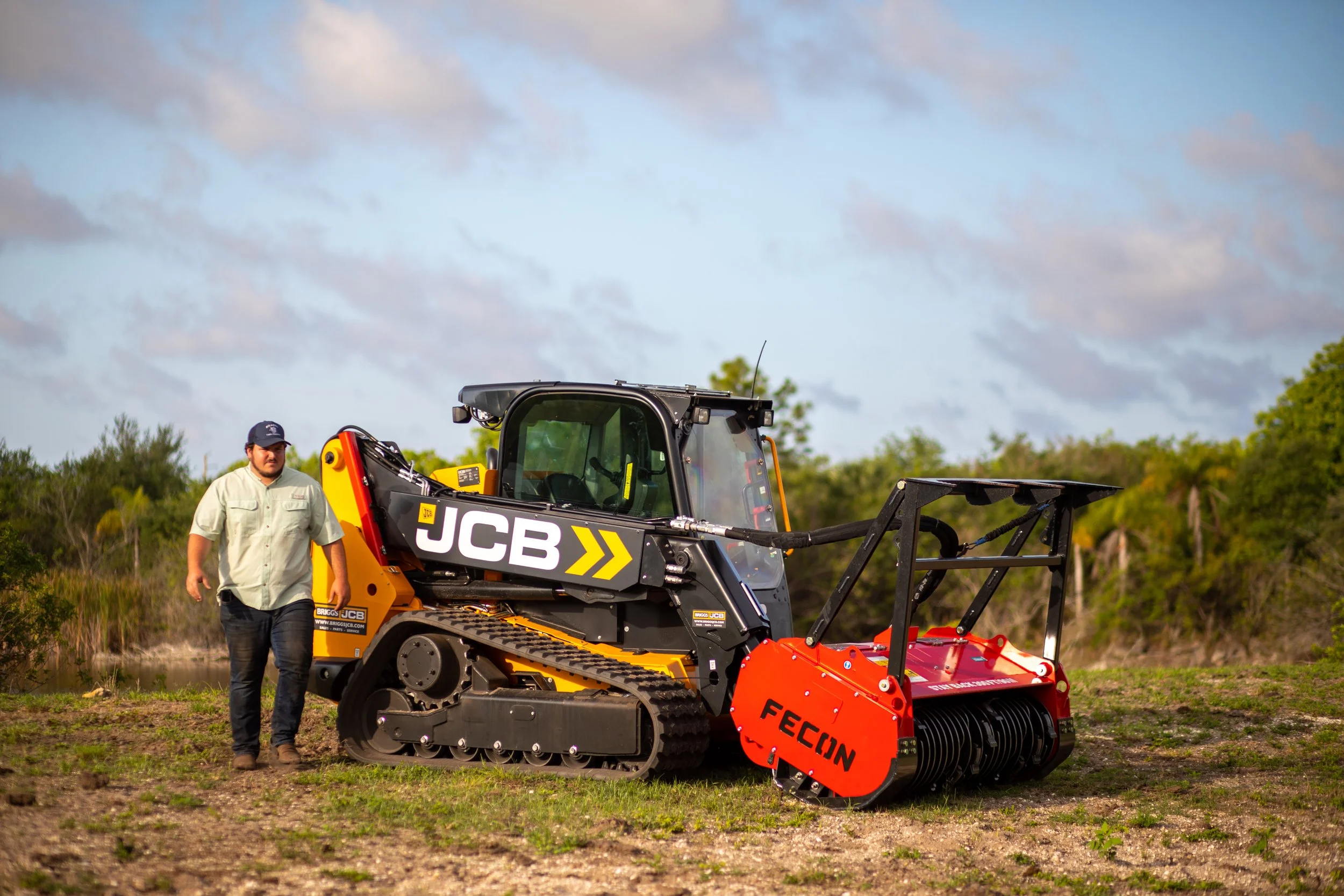 A man in a light-colored shirt and cap walking beside a JCB compact excavator with a FECON attachment on a grassy field with trees and a partly cloudy sky in the background.
