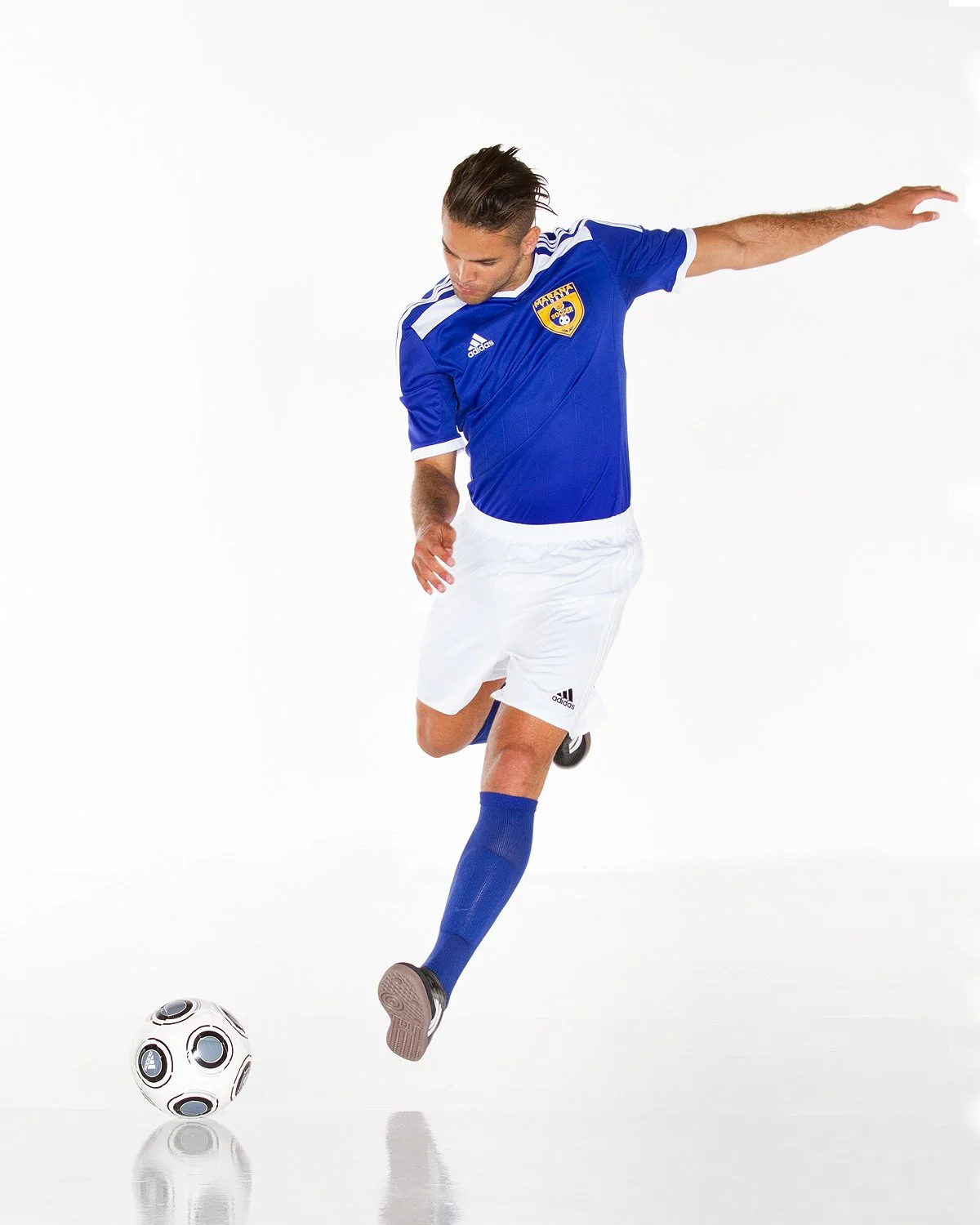 A male soccer player in a blue jersey and white shorts kicking a soccer ball indoors.