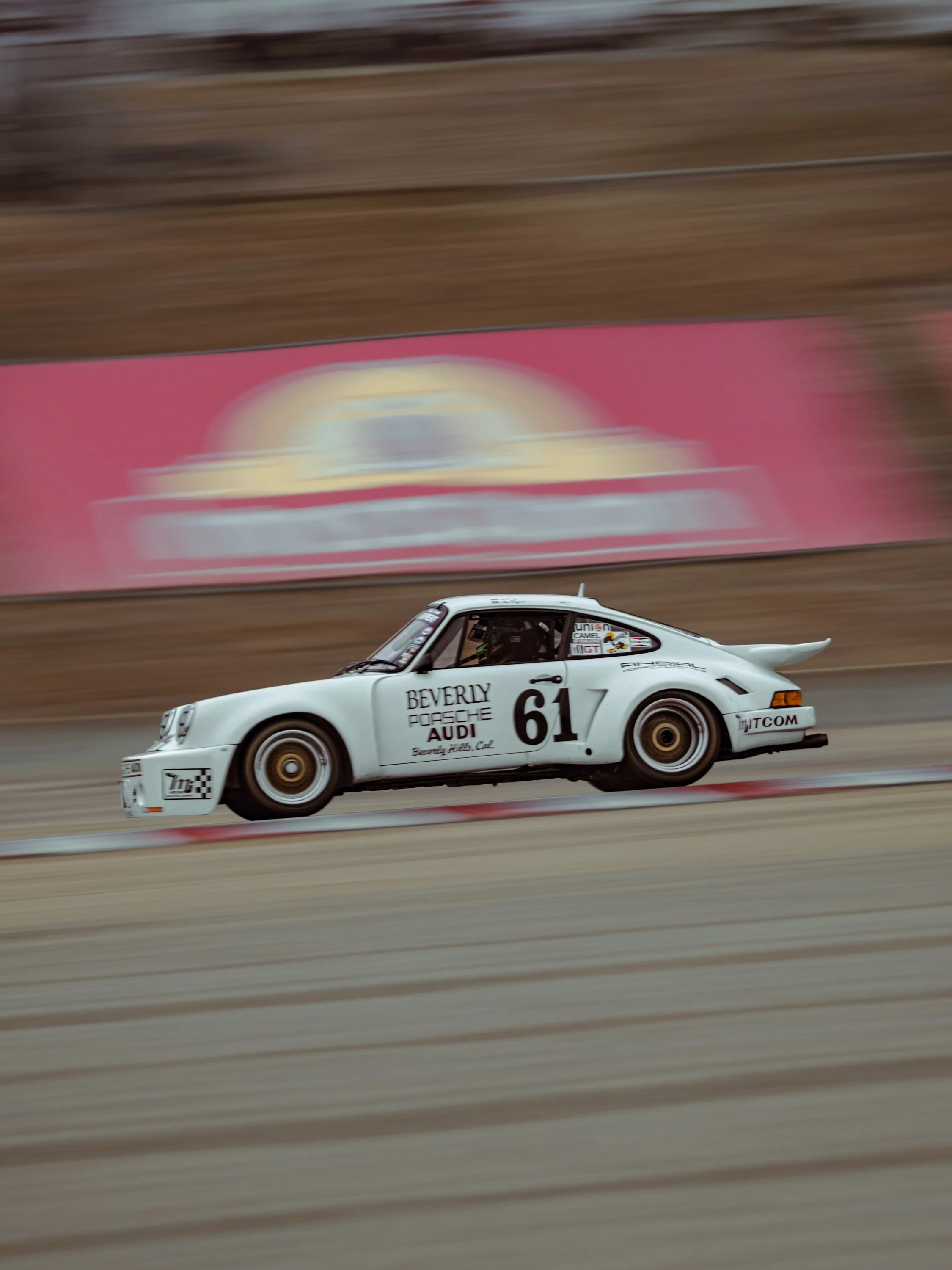 A white race car with the number 61 on its side speeding down a race track, with a blurred background of wooden barriers and advertisements.
