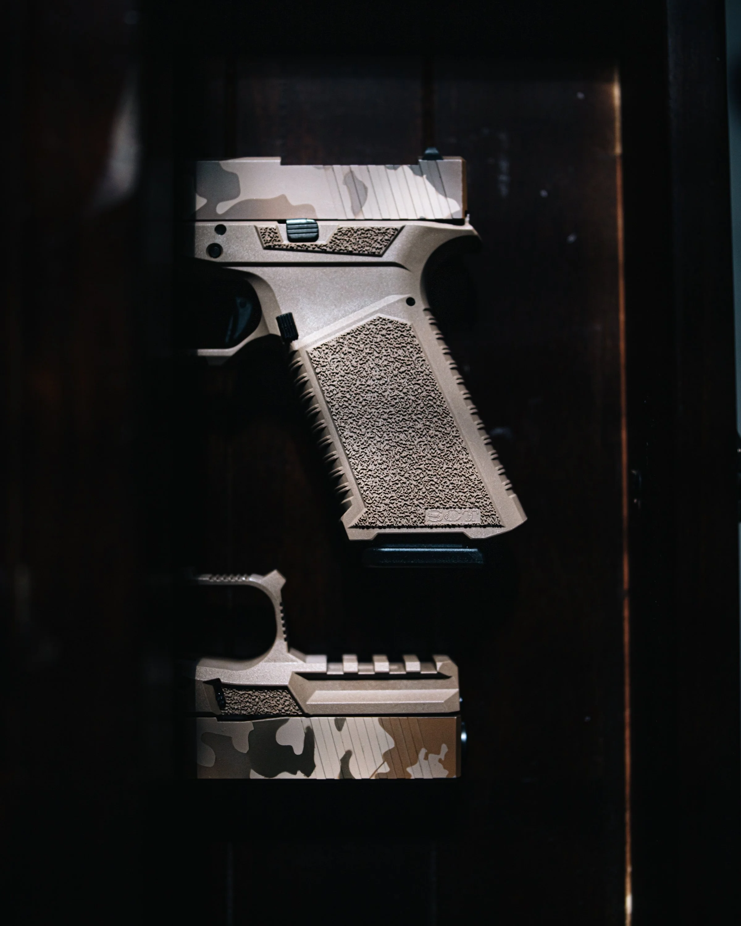 A handgun with a tan and black camo pattern is positioned vertically against a dark background, possibly inside a case or storage box. Photographed for SCT Manufacturing.