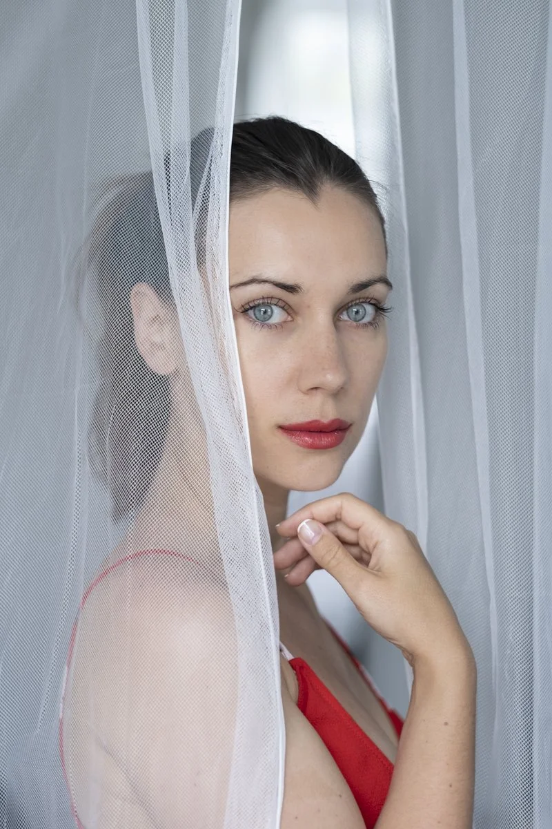 A woman with blue eyes and dark hair, wearing a red top, peeking through white sheer curtains.