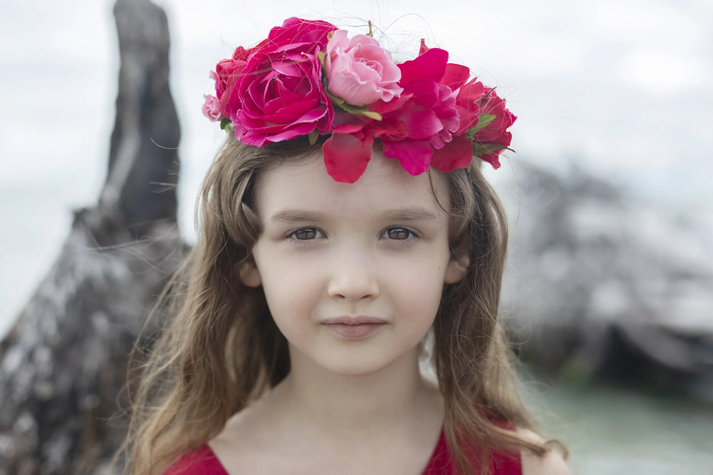 Close-up of a young girl with brown hair wearing a pink floral crown, outdoors with a blurred tree in the background.