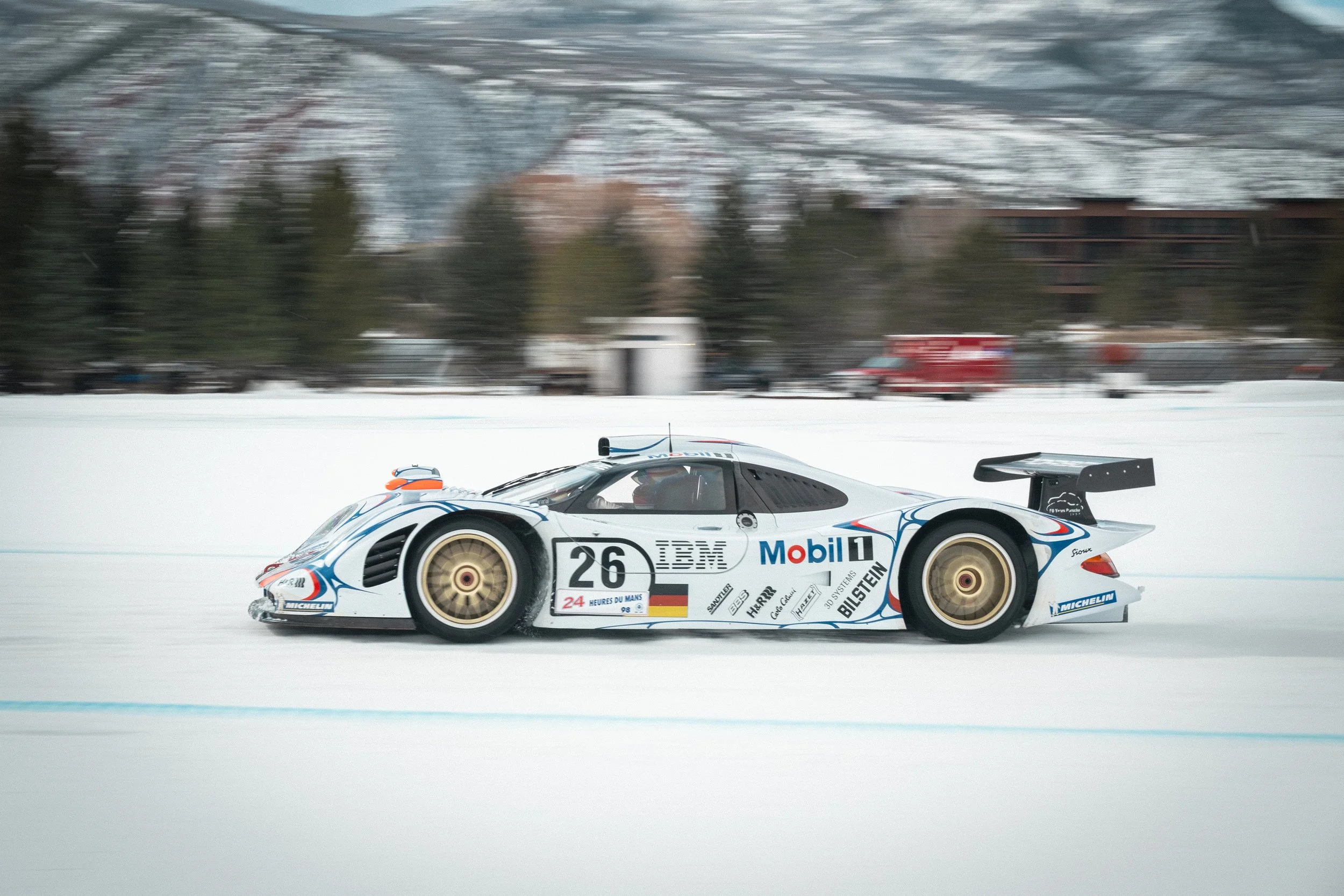 A race car moving on a snowy track with trees and buildings in the background. Porsche LeMans race car Fat Ice Race