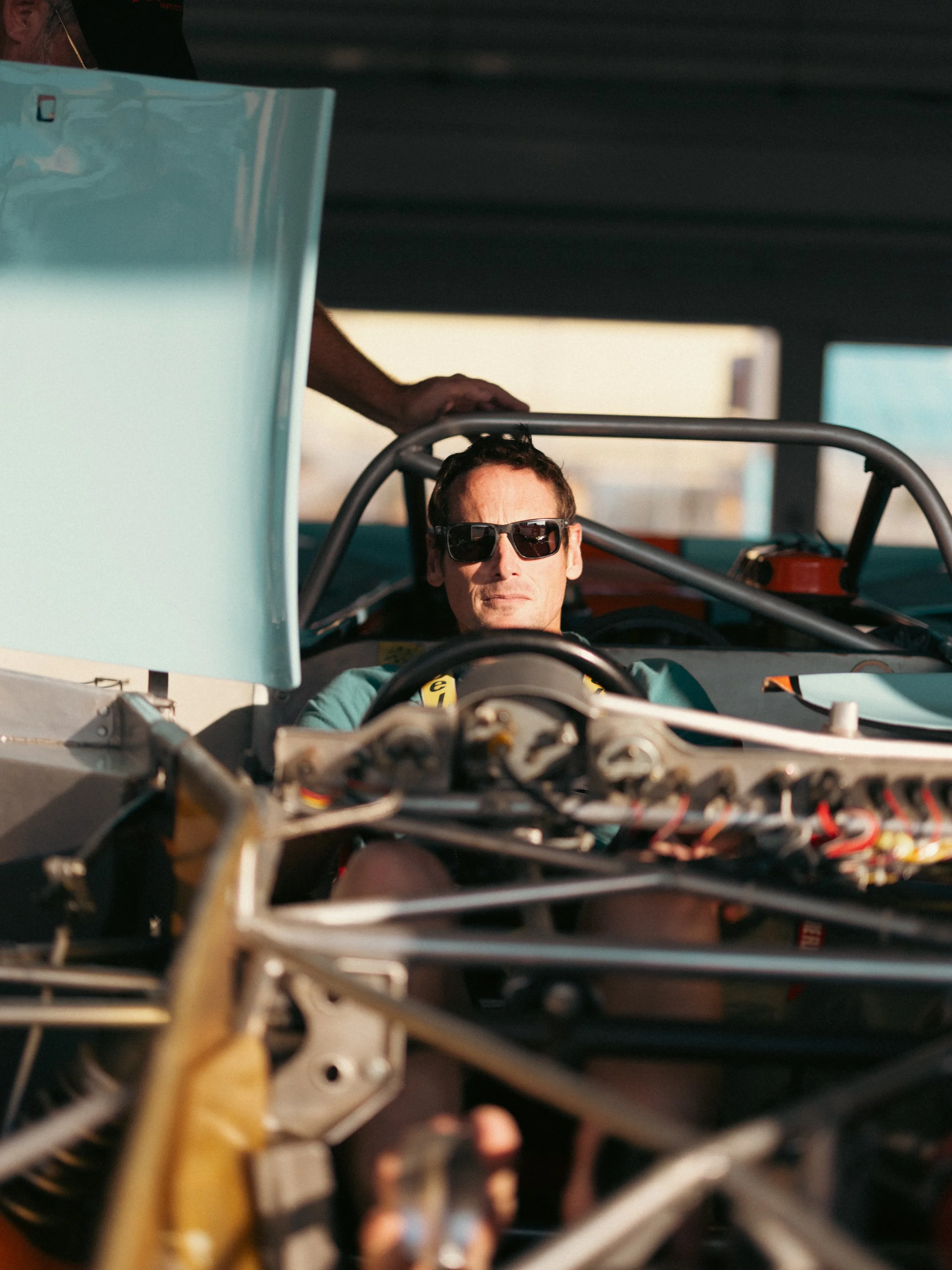 A man wearing dark sunglasses driving a race car with an open cockpit, with various wires and mechanical parts visible in the foreground, and a blurred outdoor background.