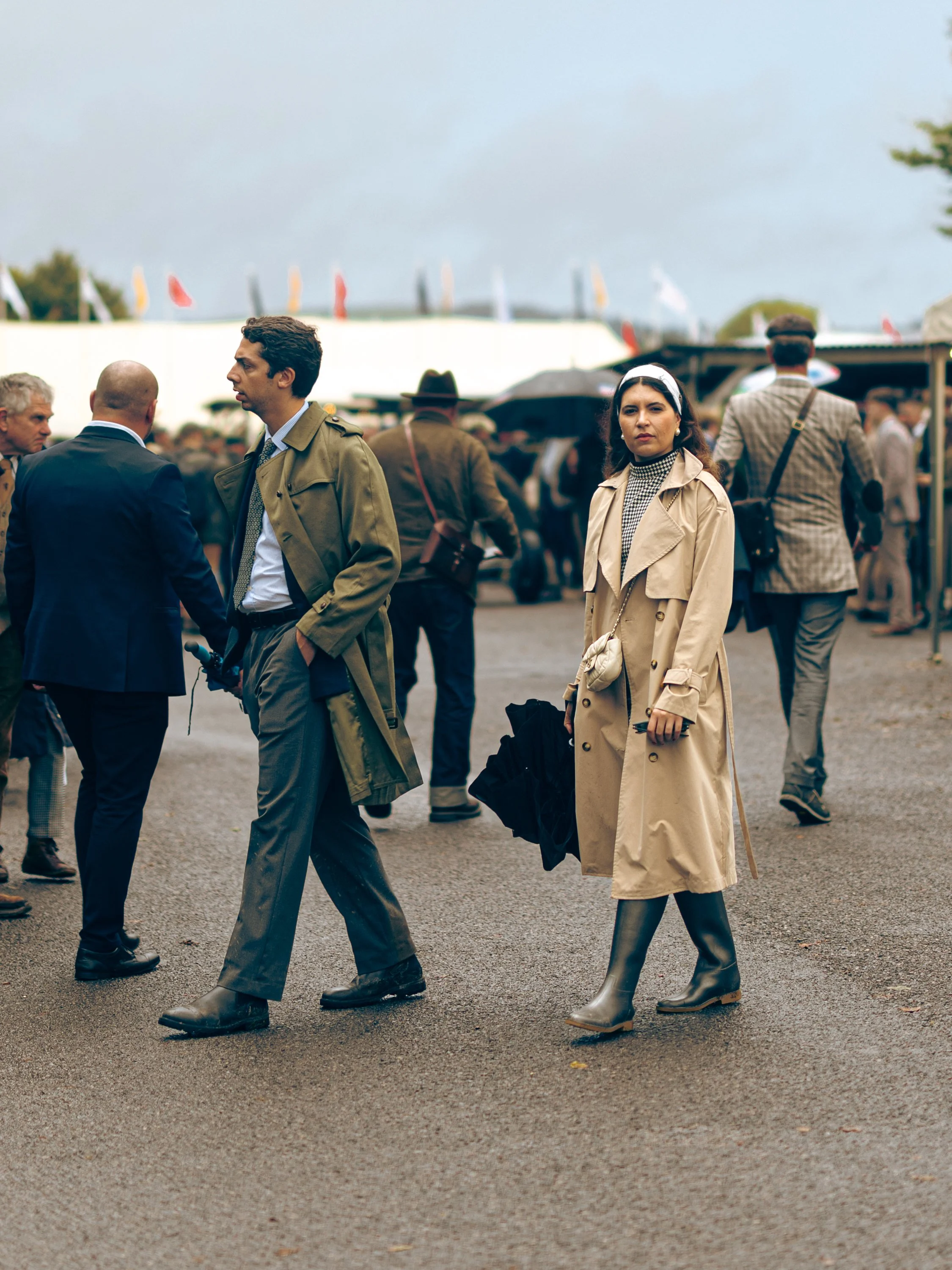 People walking outdoors on a cloudy day, with some dressed in trench coats and rain boots, and tents and flags in the background.