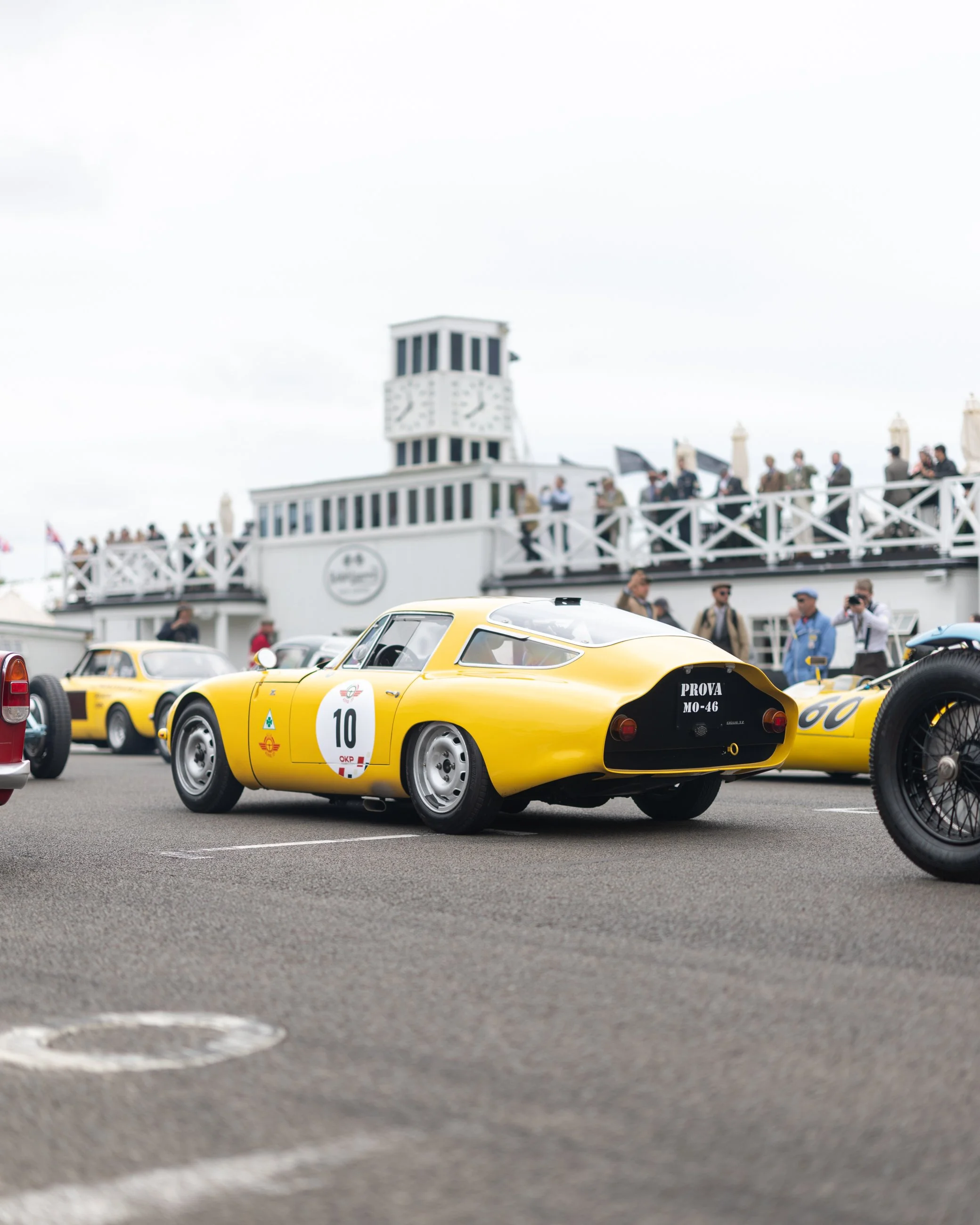 A yellow racing car with the number 10 on its side, parked on a race track, with a crowd of people and a building with a clock tower in the background. Goodwood Revival. 