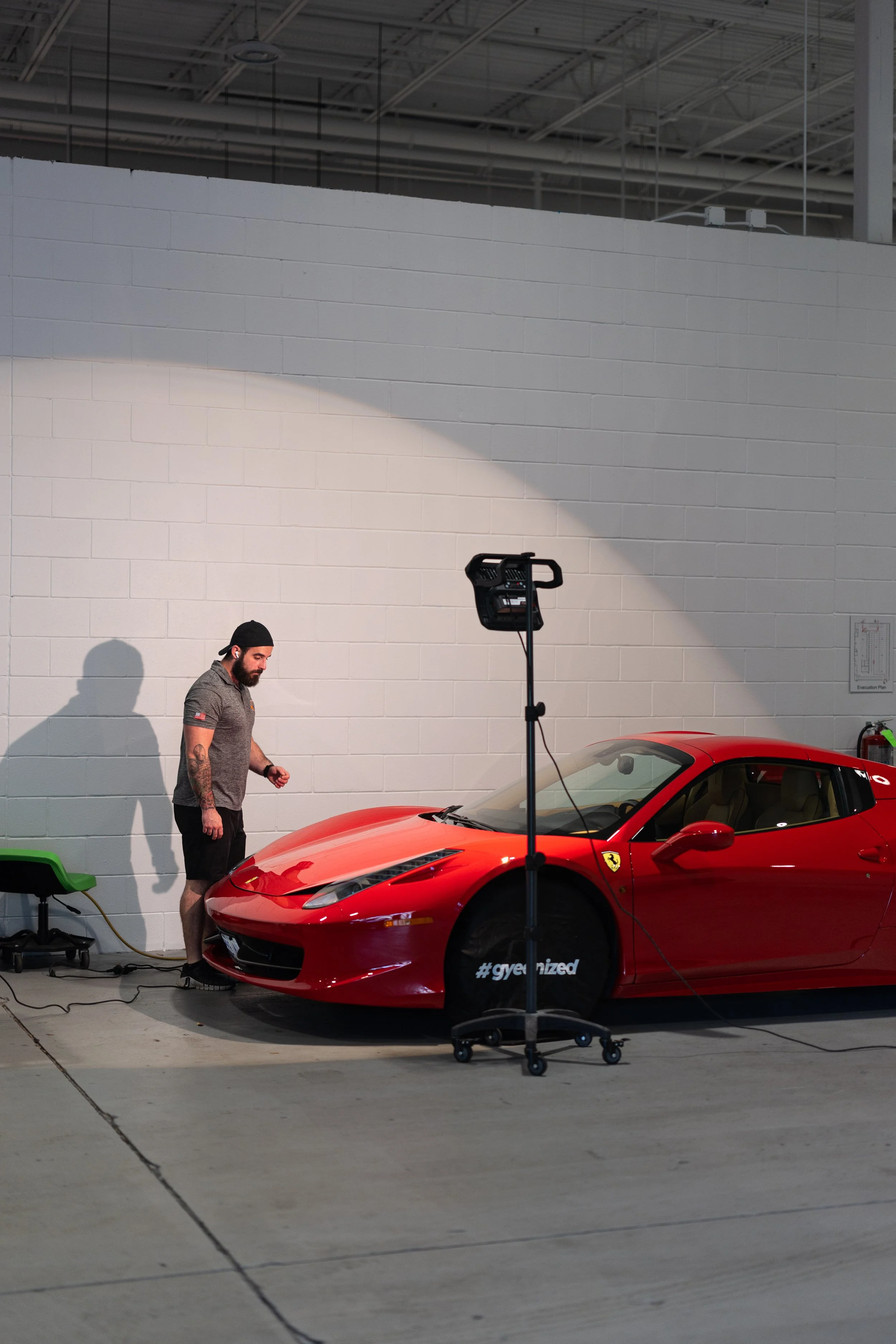 A man with tattoos and a black cap is charging a red Ferrari sports car indoors, with a white brick wall and industrial ceiling in the background. There is a light setup and some equipment nearby.