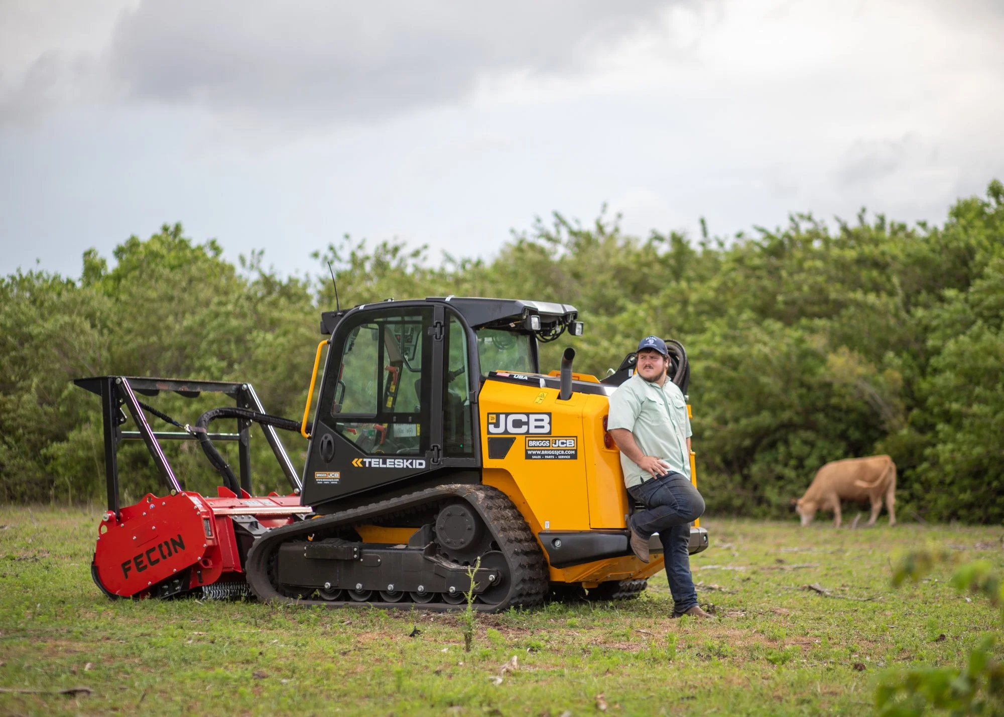 A man is standing next to a yellow and black JCB Teleskid compact tracked loader on a grassy field, with green trees and cloudy sky in the background.