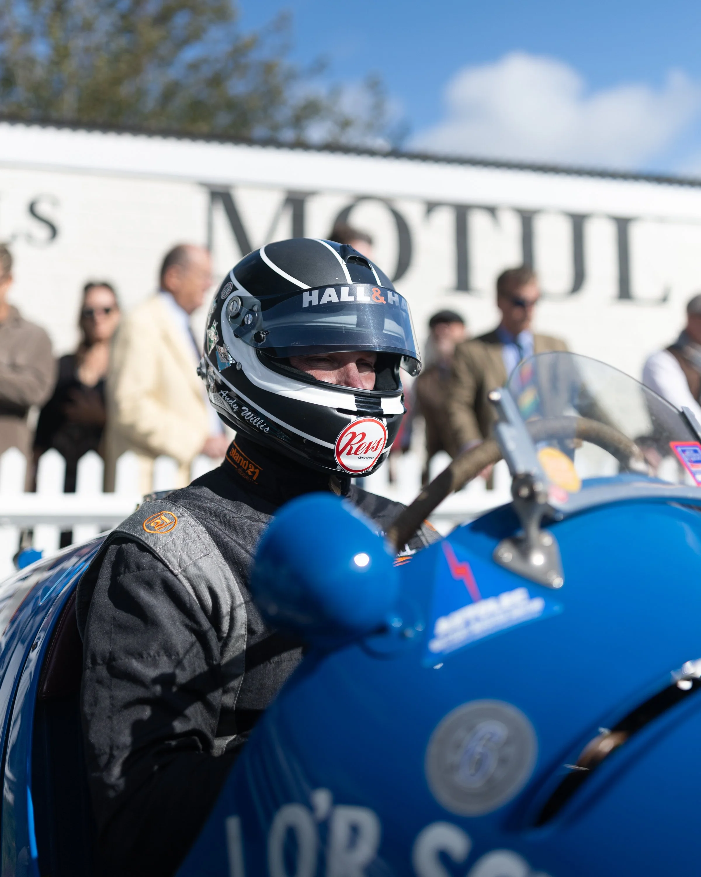 Close-up of a person wearing a racing helmet and suit, sitting on a blue motorcycle at a racing event, with spectators blurred in the background, under a clear blue sky.