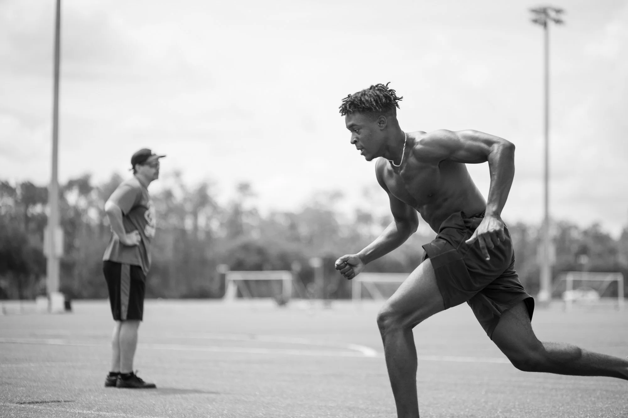 A shirtless young man with dreadlocks running on a sports field during the daytime, with a coach or trainer in the background, and tall light poles in the distance.