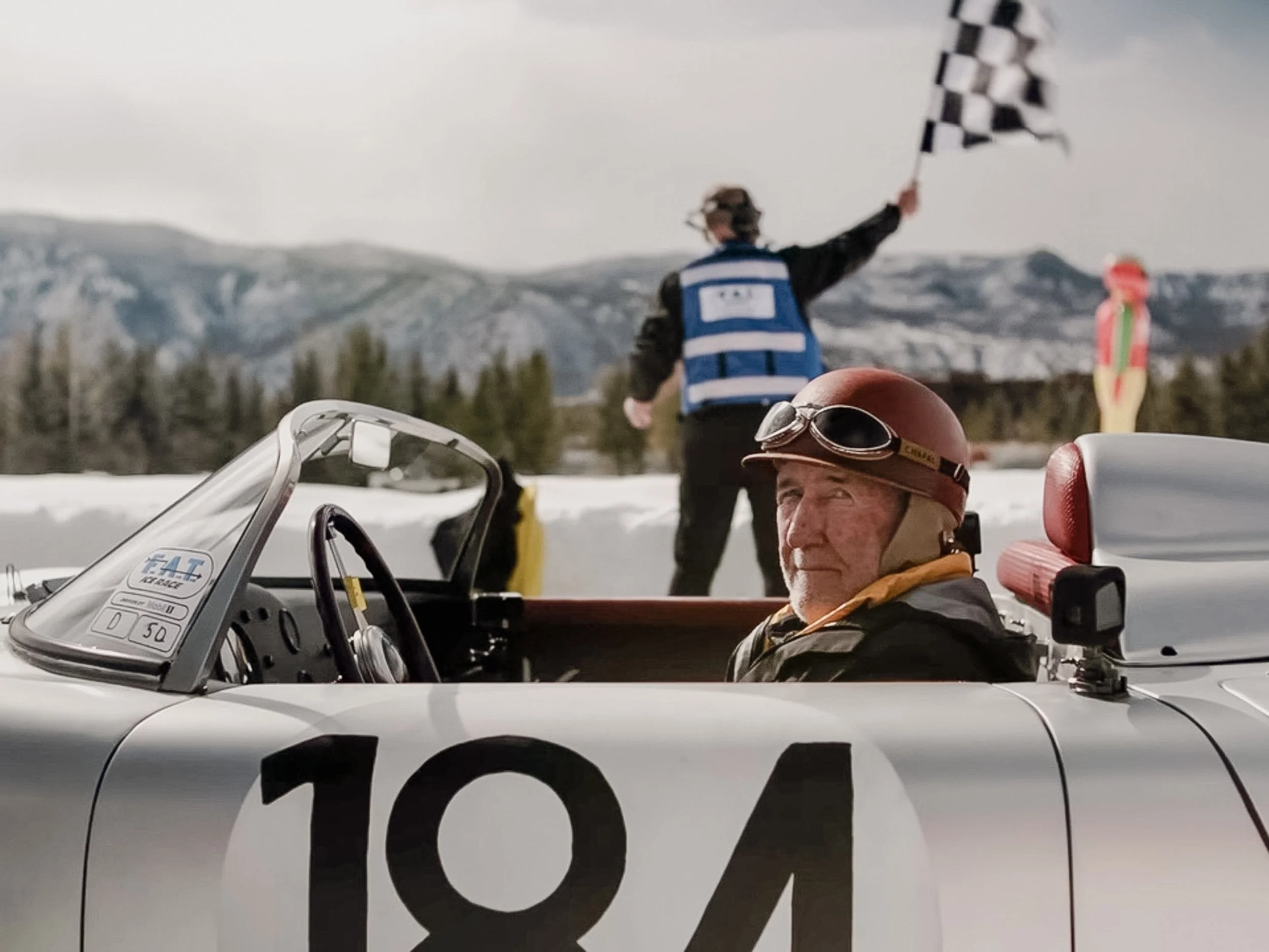 A person holding a checkered flag while standing in front of a mountainous landscape, with a motorcyclist and part of a vintage race car in the foreground. Jeff Zwart, Porsche, Fat Ice Race, Aspen, Colorado