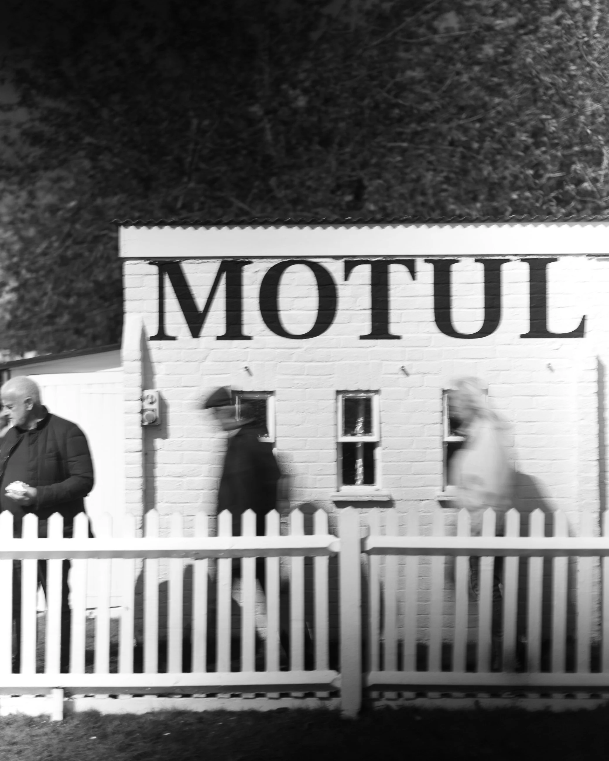 Black and white photo of a White Castle restaurant with a sign that reads 'MOTUL,' while three people walk in front of it, and a white picket fence is in the foreground. Goodwood Revival.